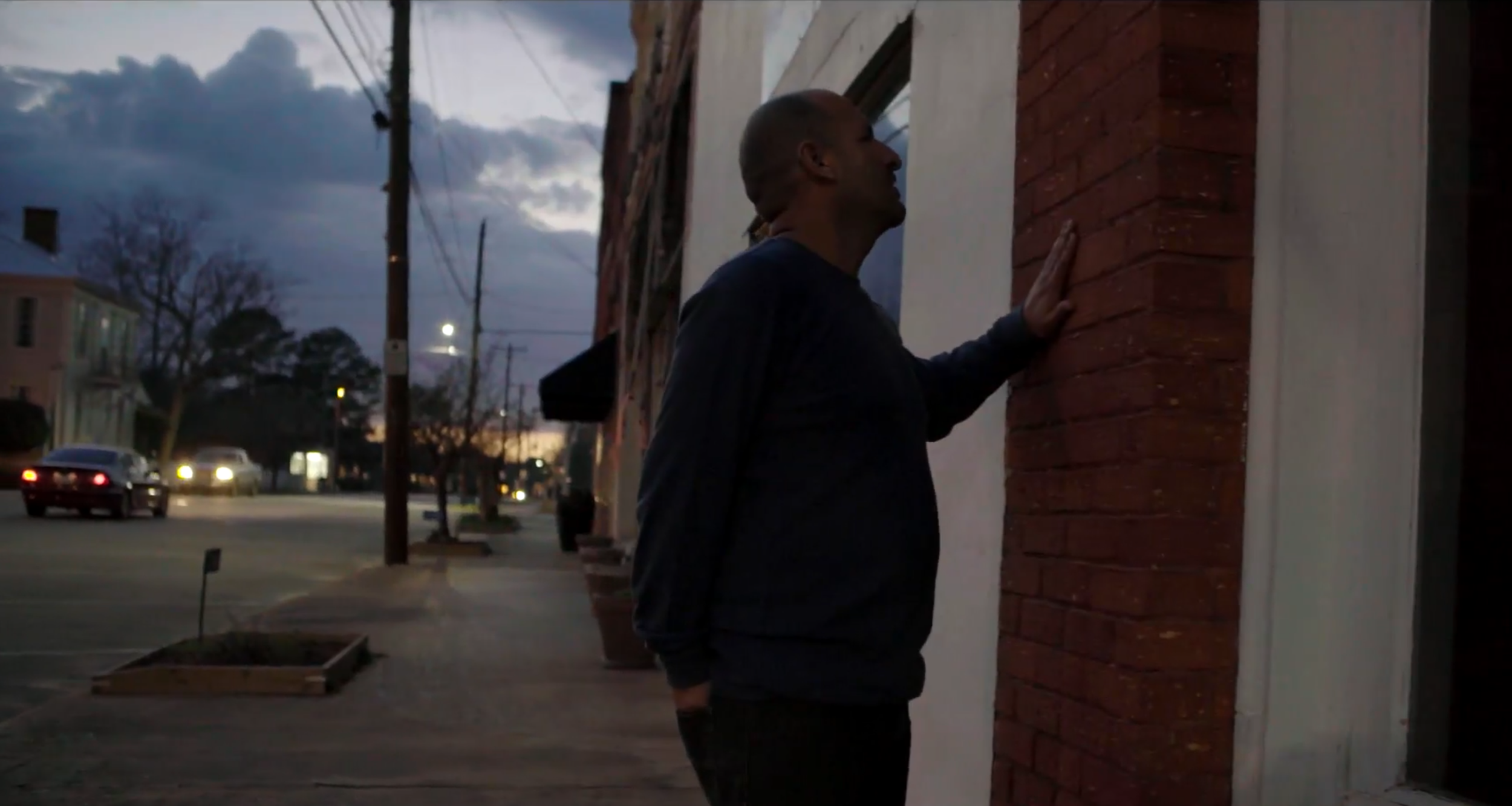 At twilight, a hispanic man places his hand against a brick wall on a small town main street