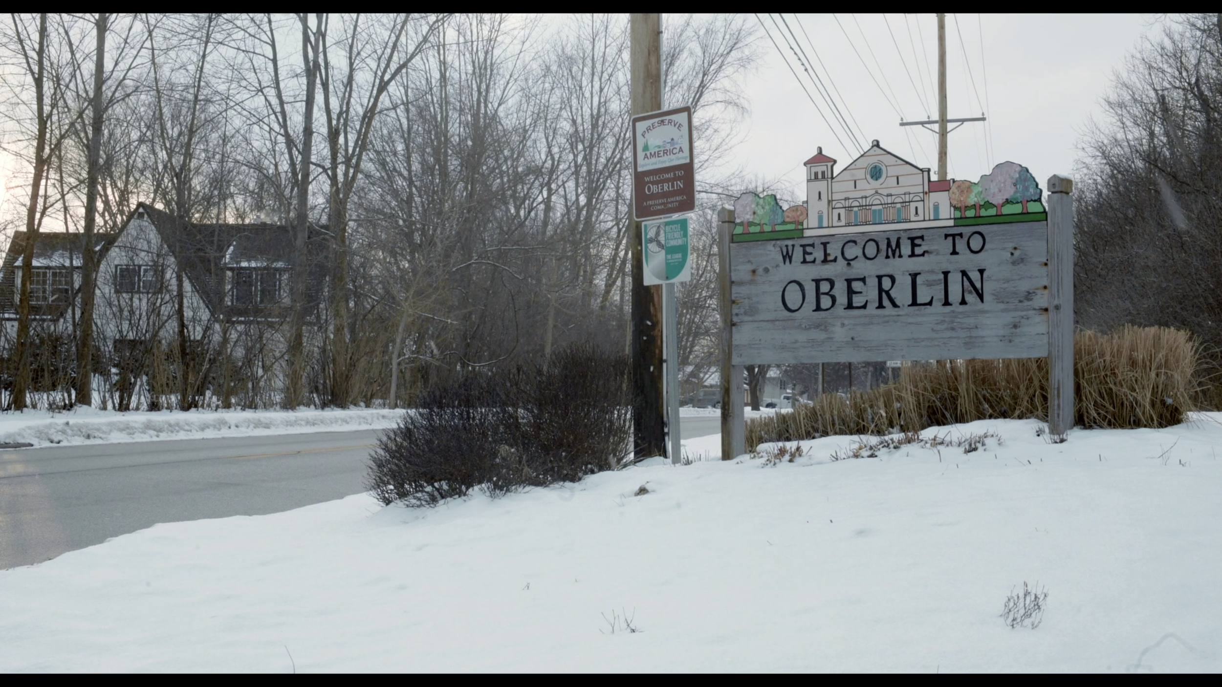 Amidst a snow covered landscape, a sign beside a narrow road reads WELCOME TO OBERLIN