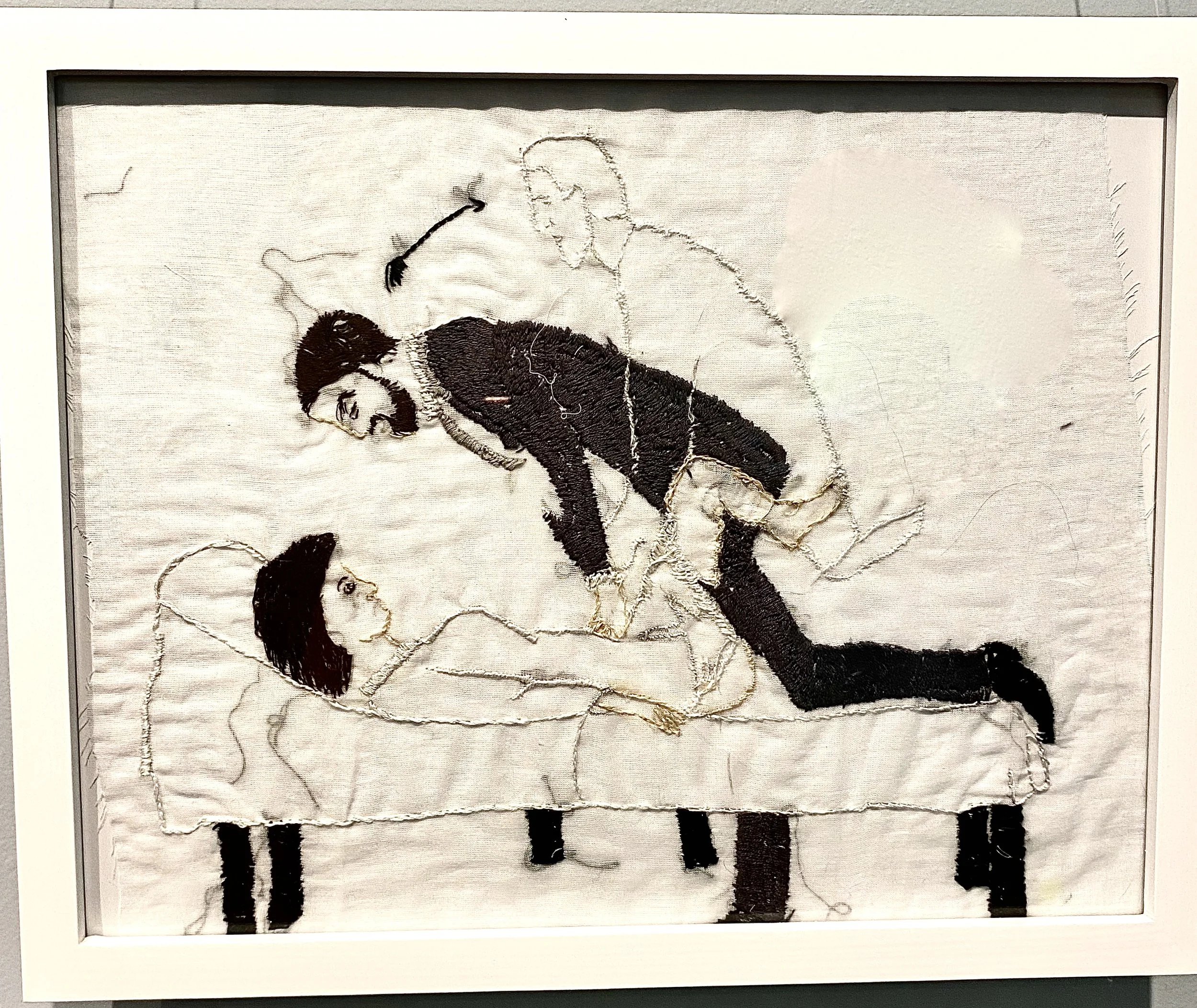 Embroidered art depicting three figures, one on a bed and two standing. The artwork features prominent use of black, white, and brown threads, creating a textured visual effect.
