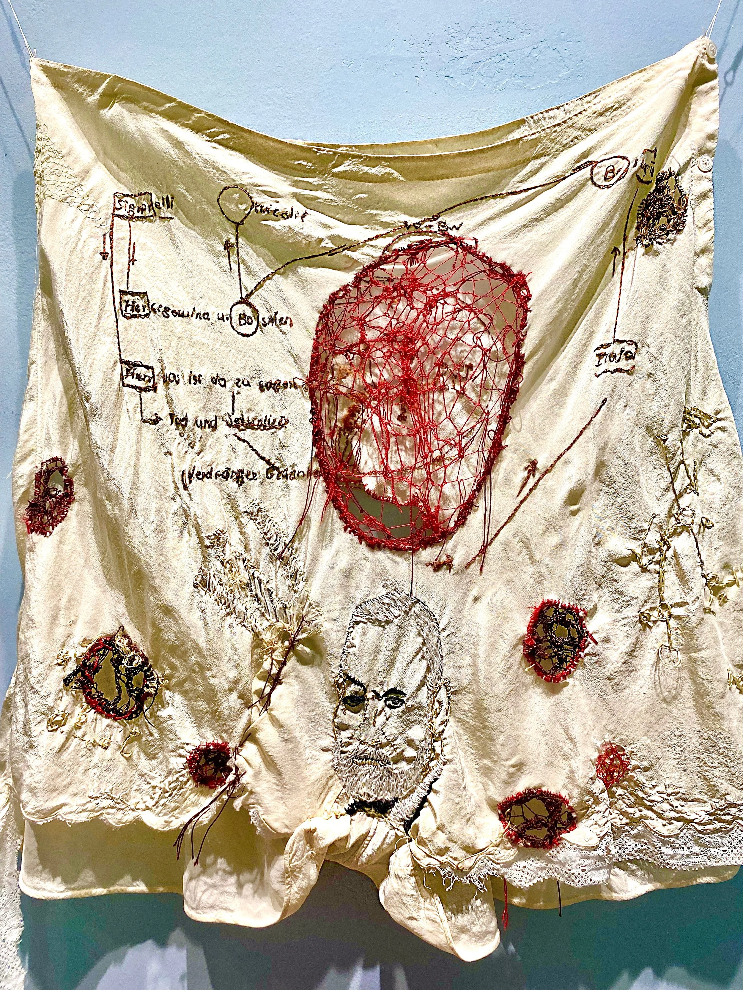 An embroidered textile artwork featuring abstract designs and text, with a prominent red stitched pattern in the center. There is a portrait of a man with a beard stitched in black and white thread. Other elements include red circular patterns and lines, all on a beige fabric background.