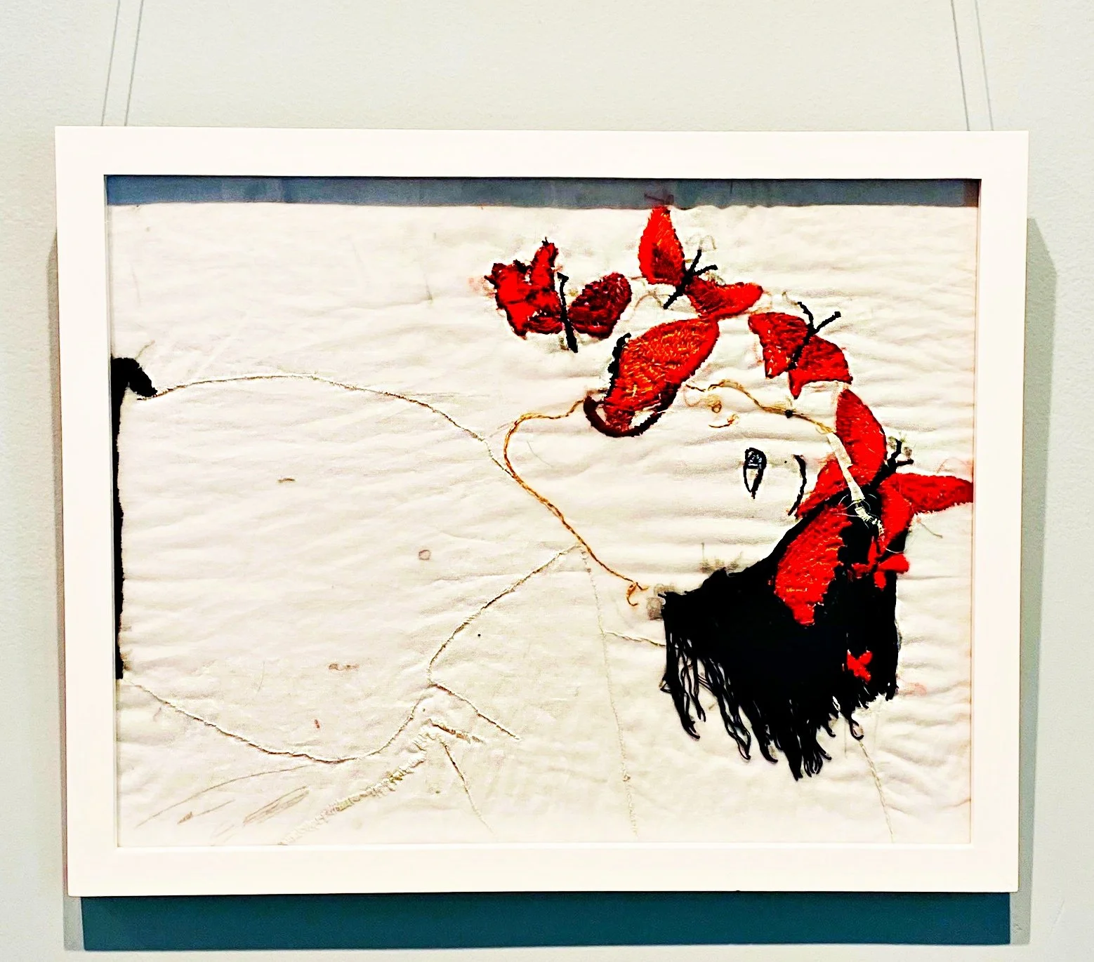 Abstract embroidery artwork with red and black thread on white fabric, depicting a face silhouette and red leaves or petals.