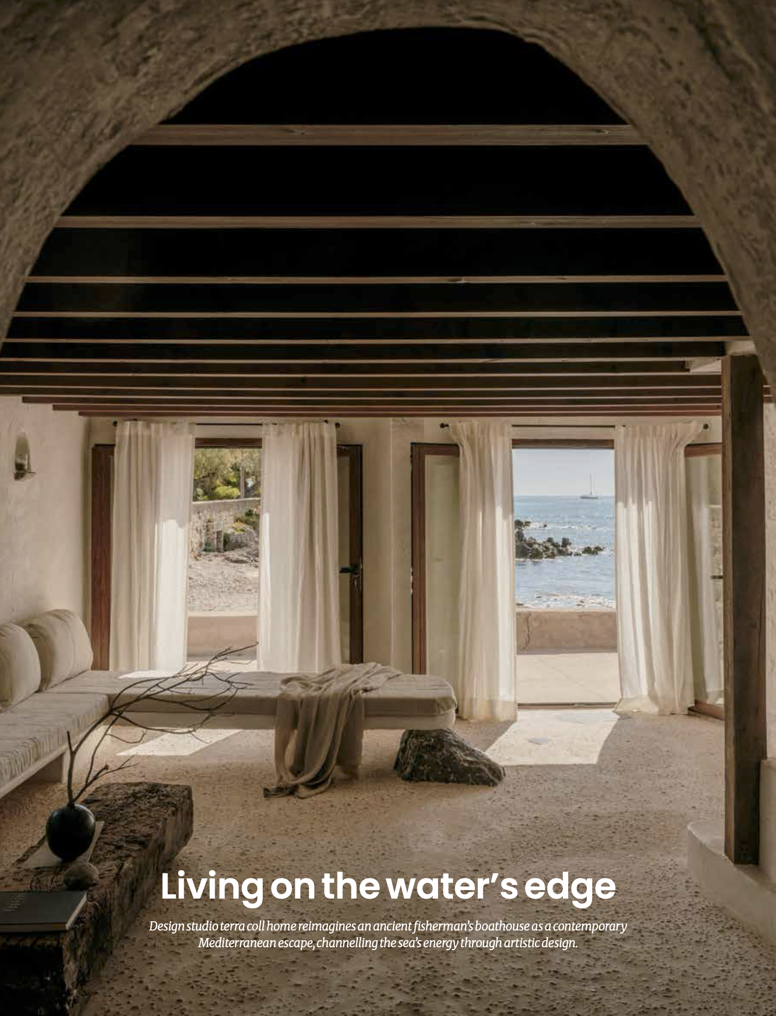 Interior of a beach house with large windows showing the ocean and sky outside, white curtains, a sofa, a coffee table, and decorative elements, with a view of the water's edge.