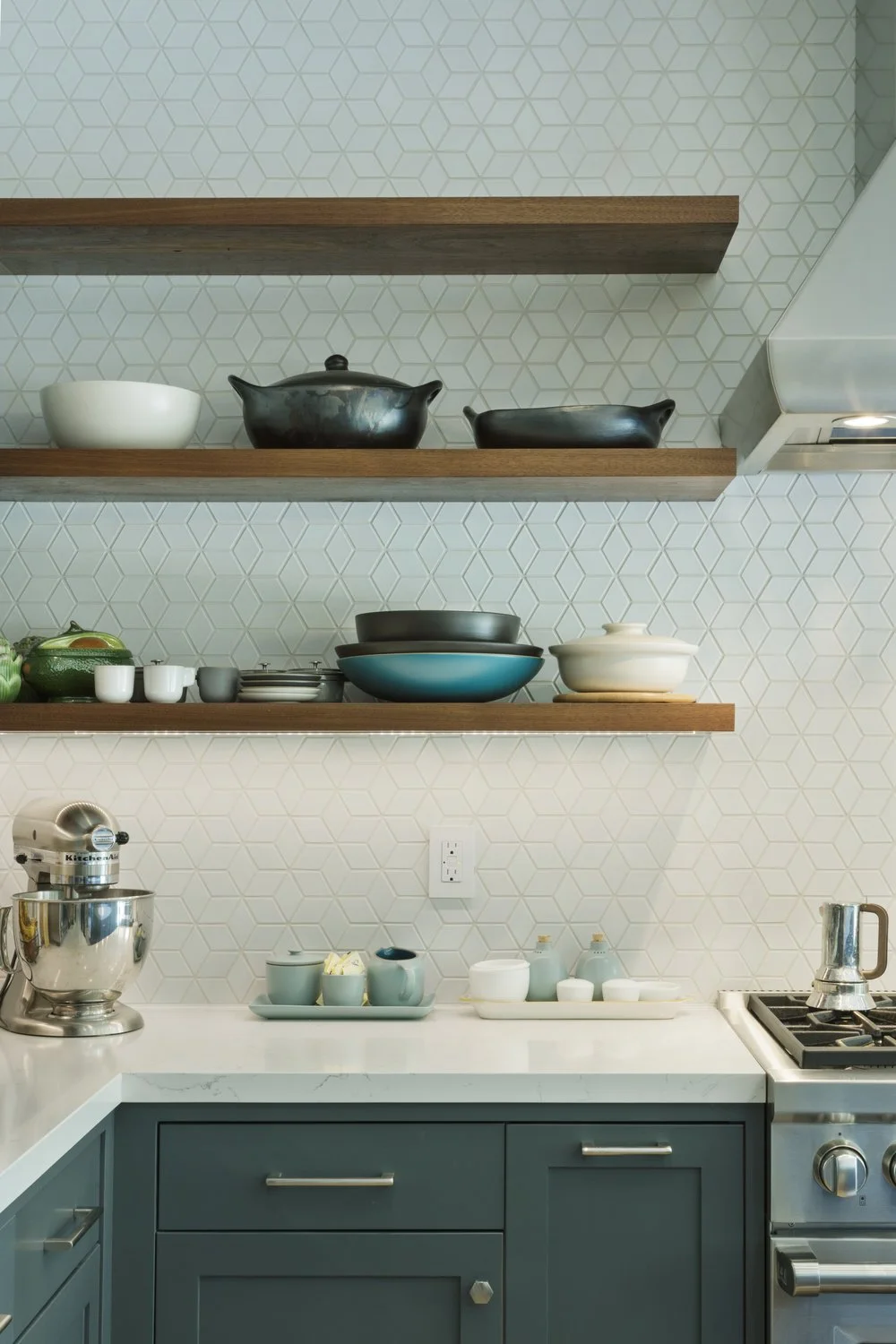 Kitchen with open wooden shelves holding black, white, and ceramic bowls and dishes. Blue cabinetry with silver handles, a white marble countertop, and modern appliances including a stand mixer and stovetop. White geometric patterned wall background.