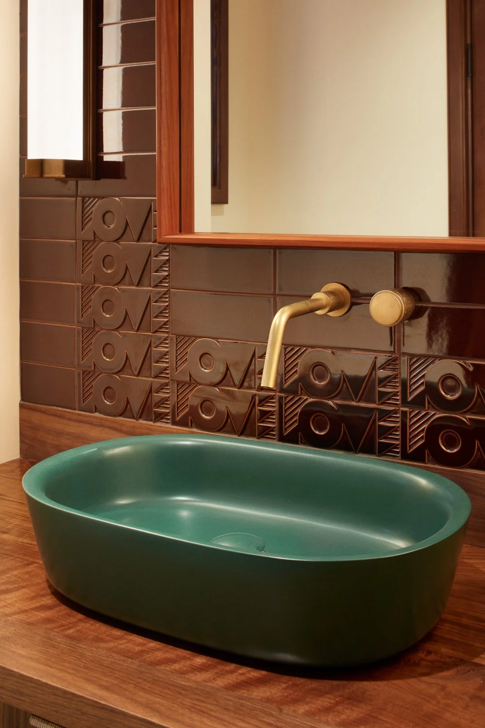 A green oval-shaped ceramic sink with a gold faucet, mounted on a wooden countertop, reflected in a wooden framed mirror against a tiled wall with geometric patterns.