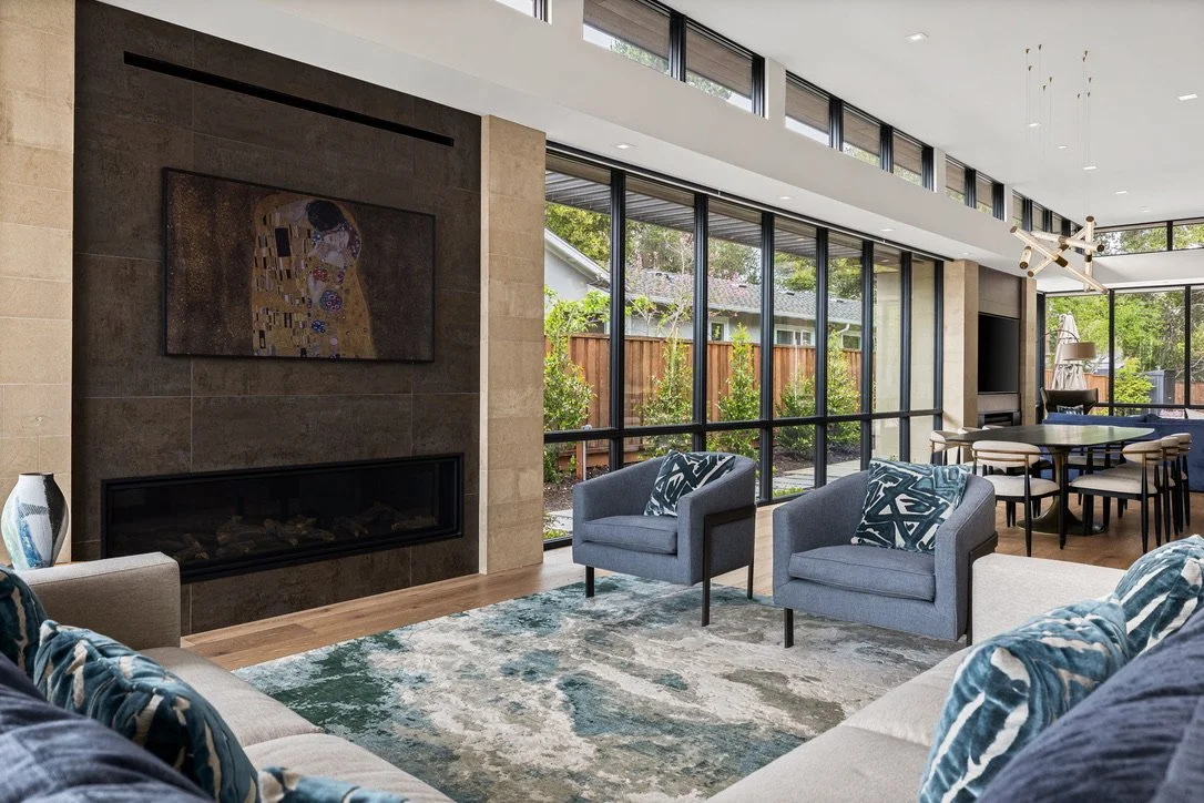 Modern living room with floor-to-ceiling windows, contemporary furniture, a fireplace with a colorful artwork above, and a view of the outdoor garden.
