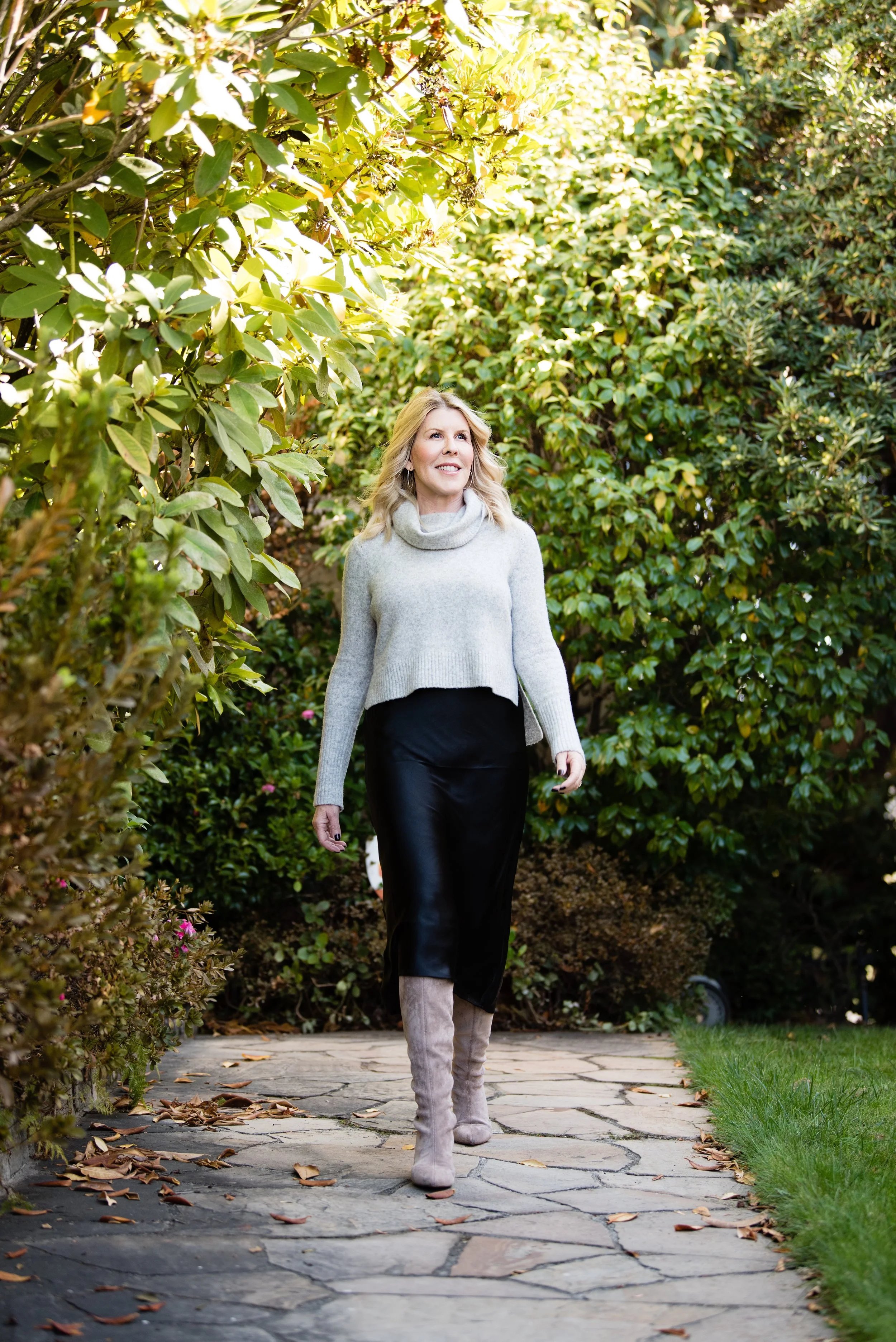 Lydia Lyons on a stone pathway in a garden, surrounded by green bushes and trees, wearing a light gray sweater, black skirt, and knee-high beige boots. Luxury Residential Interior designer in California.
