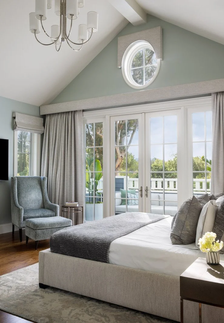 A bedroom with a bed, gray bedspread, pillows, a gray armchair, small side table, and glass doors leading to a patio with outdoor furniture and trees visible outside.