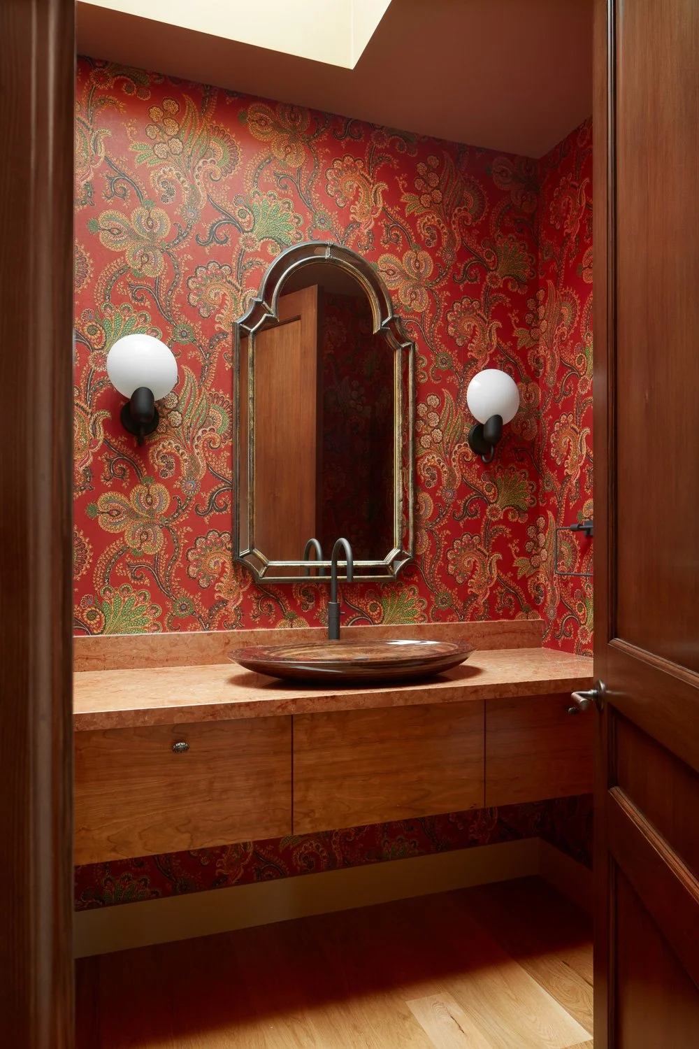 A bathroom vanity with a round vessel sink, a mirror with a decorative frame, two wall sconces with globular light bulbs, and red and gold patterned wallpaper.