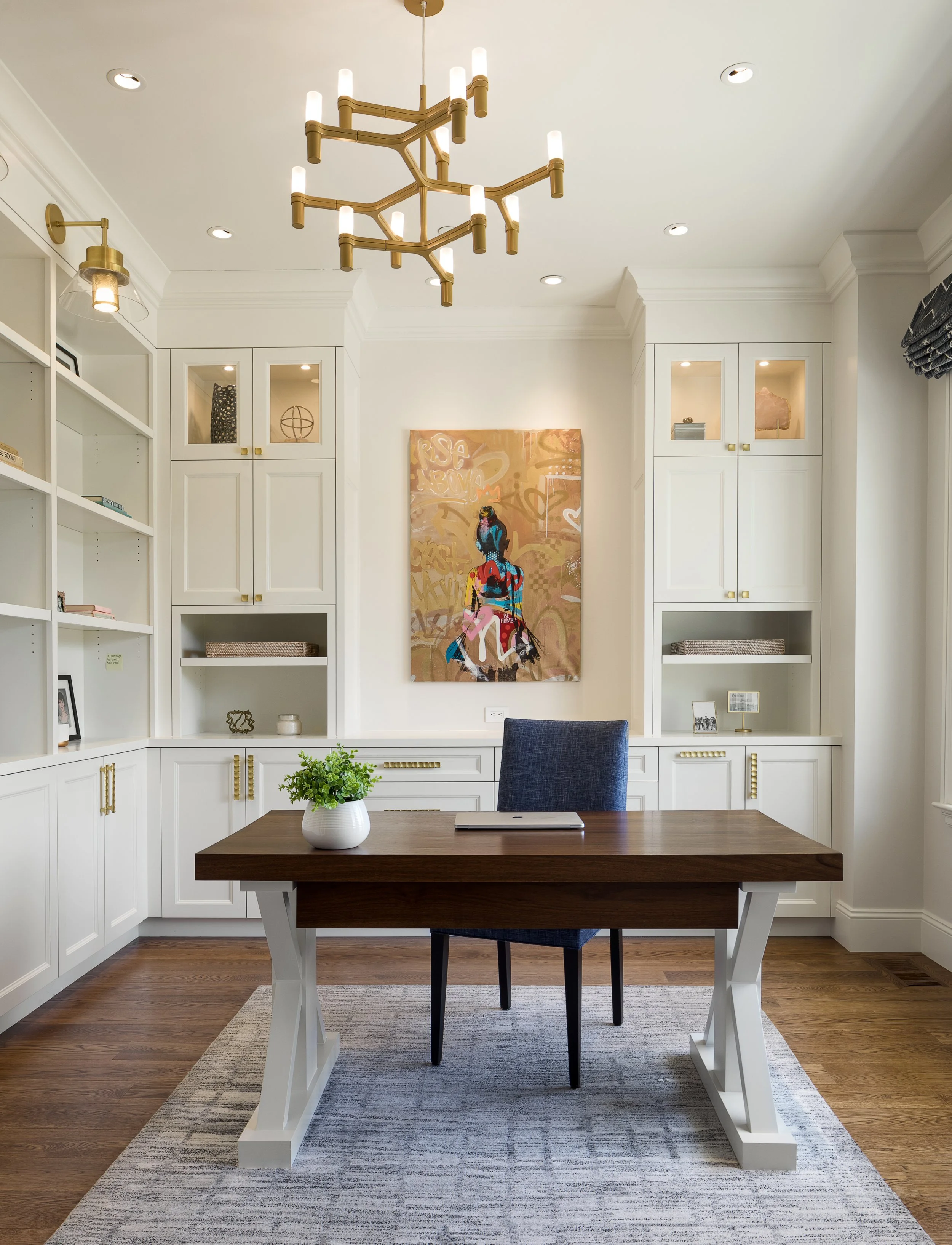 Modern dining room with white built-in shelves, black and white artwork, a wooden table with a white base, a blue upholstered chair, a potted plant, a large abstract painting, and elegant lighting fixtures.