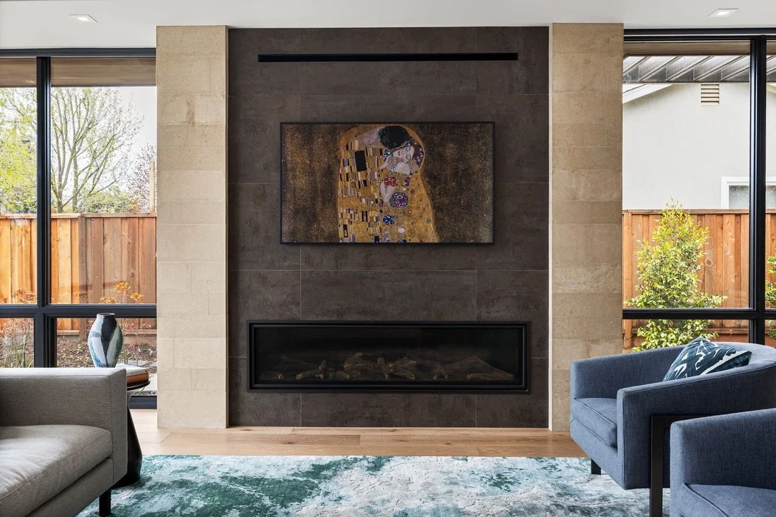 Living room interior with a modern fireplace, framed artwork above, large windows with views of a fenced backyard and trees, and blue and beige seating.