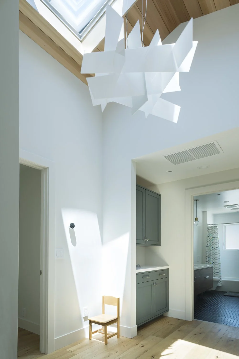 Modern bathroom with a skylight, a hanging white geometric light fixture, a small wooden chair, gray cabinetry, and a bathroom with a shower curtain and sink.