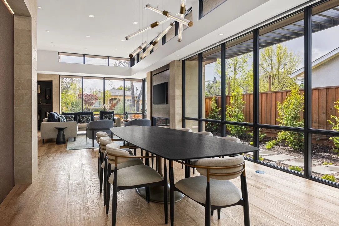 Modern living room and dining area with large windows, wooden floors, and contemporary lighting fixture.