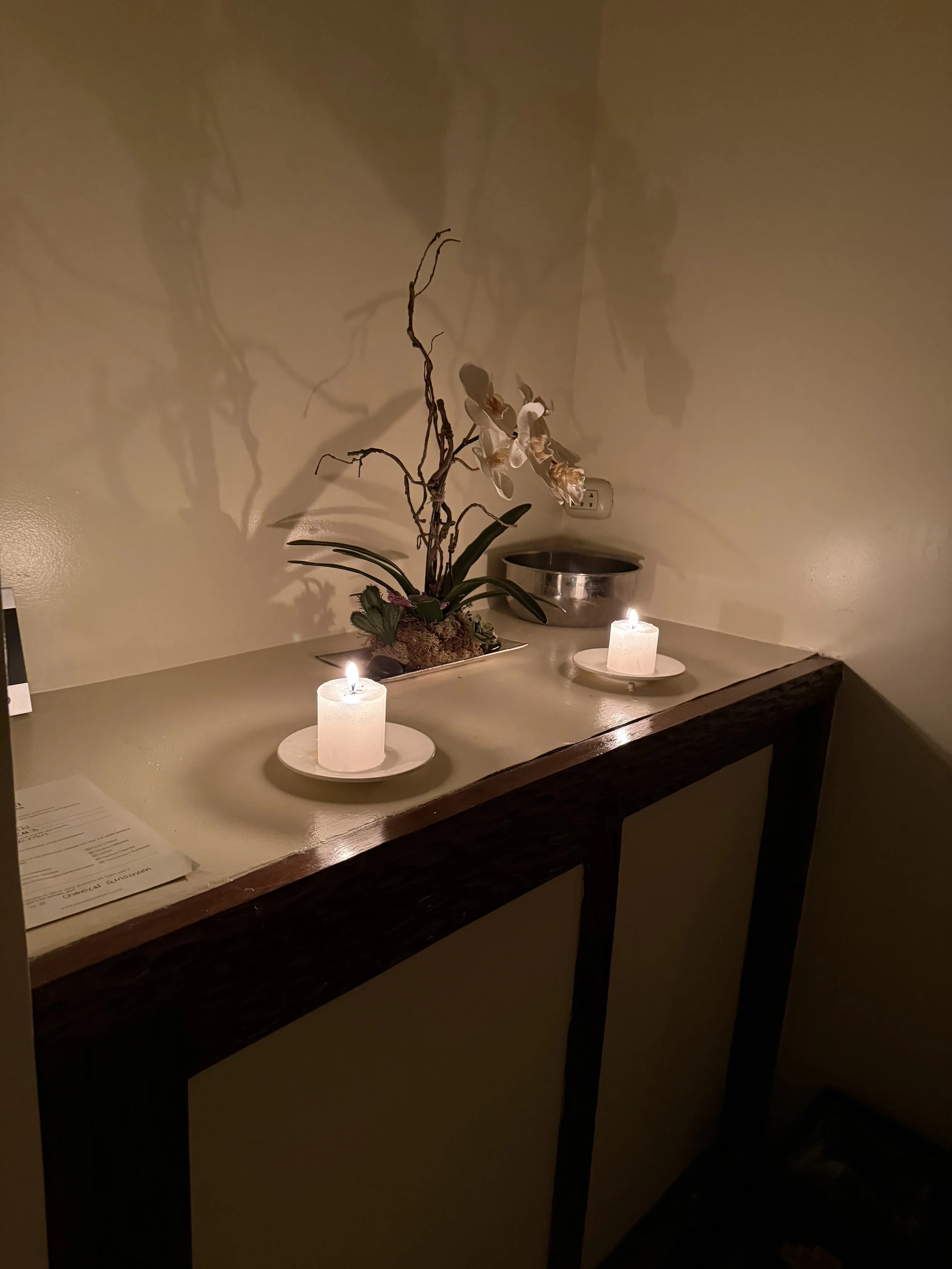 beige console displaying two white candles with wicks burning.  a floral arrangement placed behind the candles.