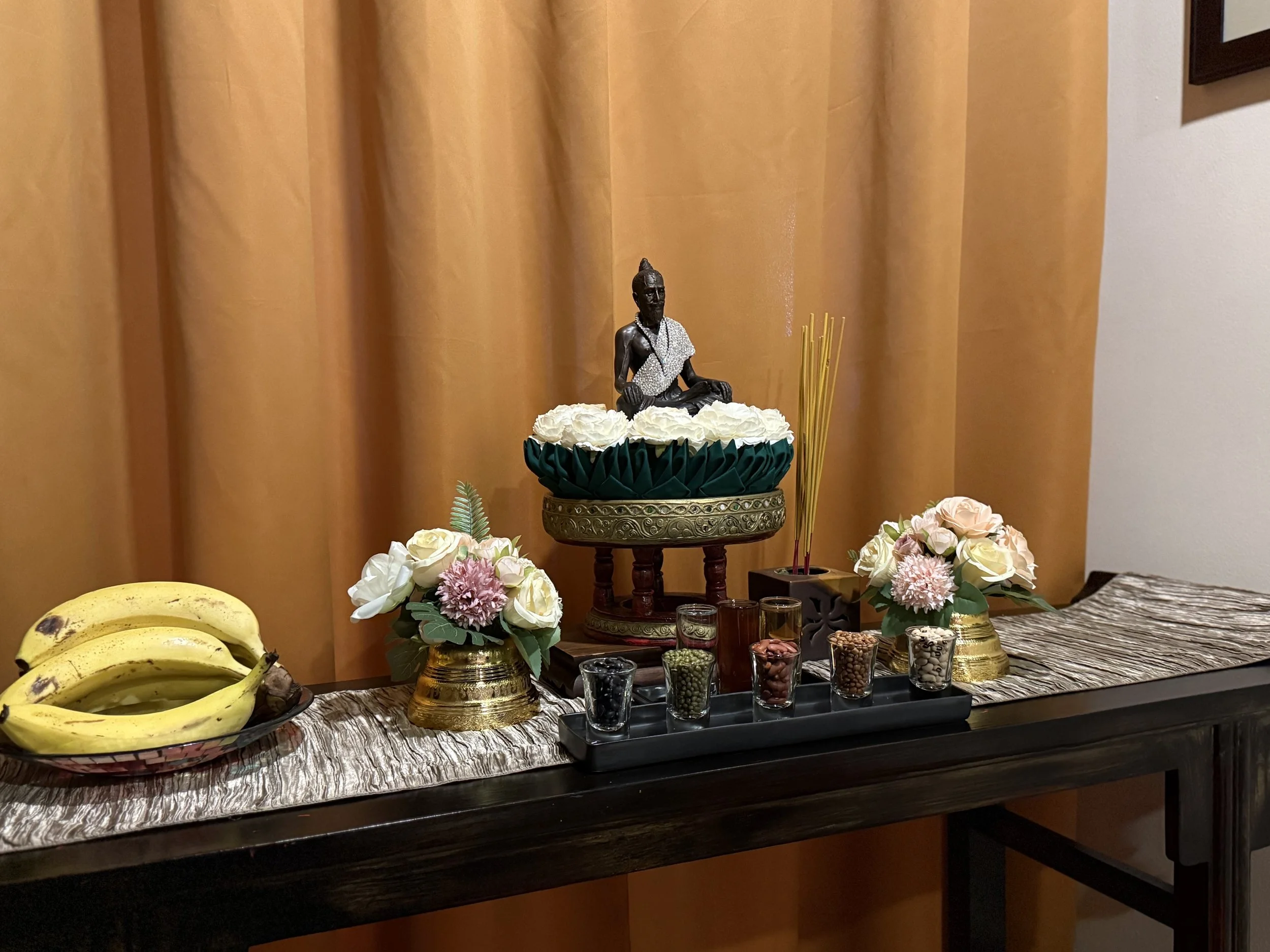 A long narrow table with an image of a buddha placed on a pedestal and surrounded by flowers.  Two floral arrangements flank the pedestal.  A long tray is in front of the pedestal.  Five small glasses are in tray.  Each holds a type of beans