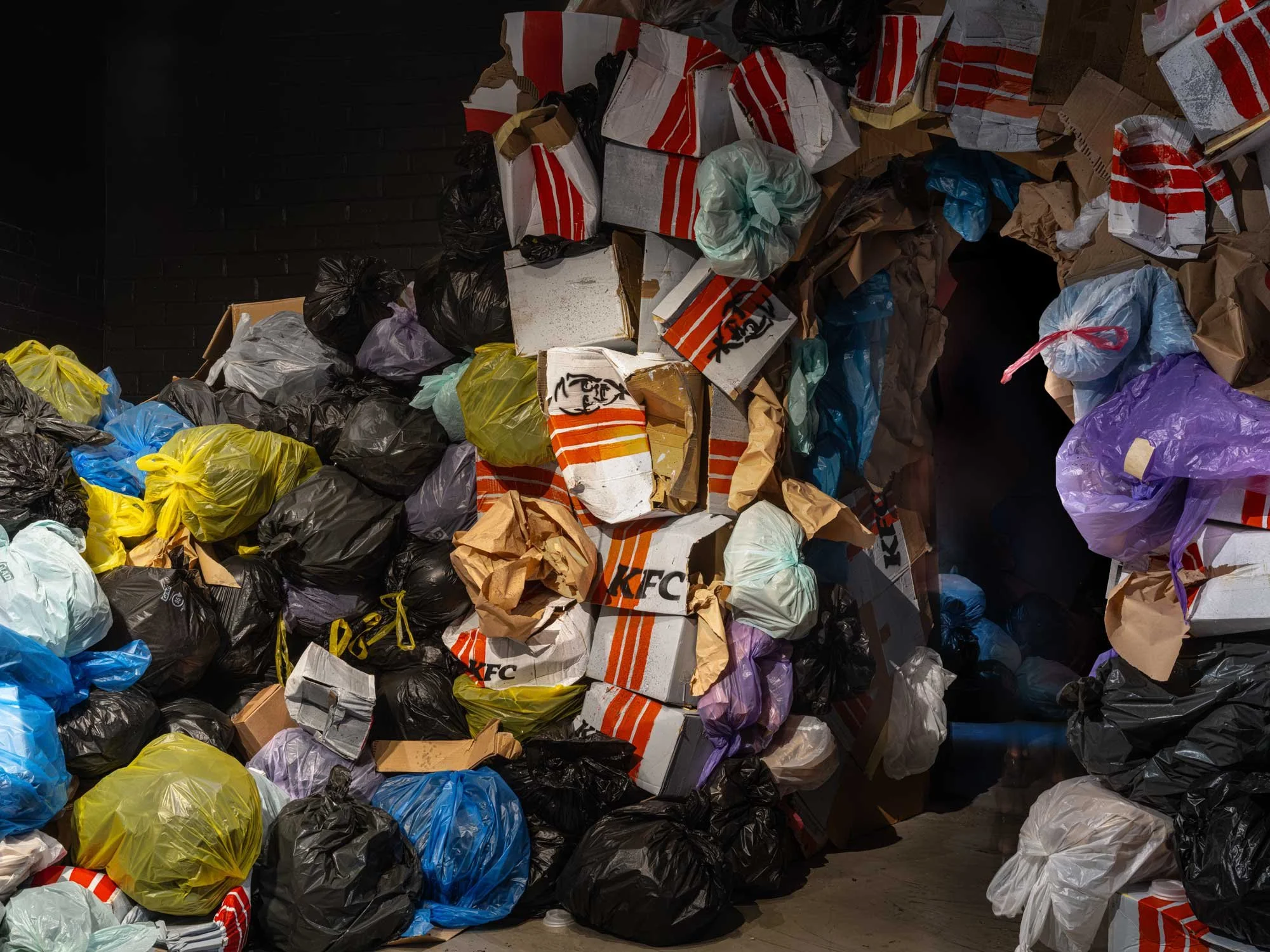 A large pile of trash bags and cardboard boxes, including KFC pizza boxes, in a dark indoor setting. Mysterious al immersive pop up art exhibition Human Nature, Art in disused warehouse, pop-up spaces, colourful, striking contemporary art.