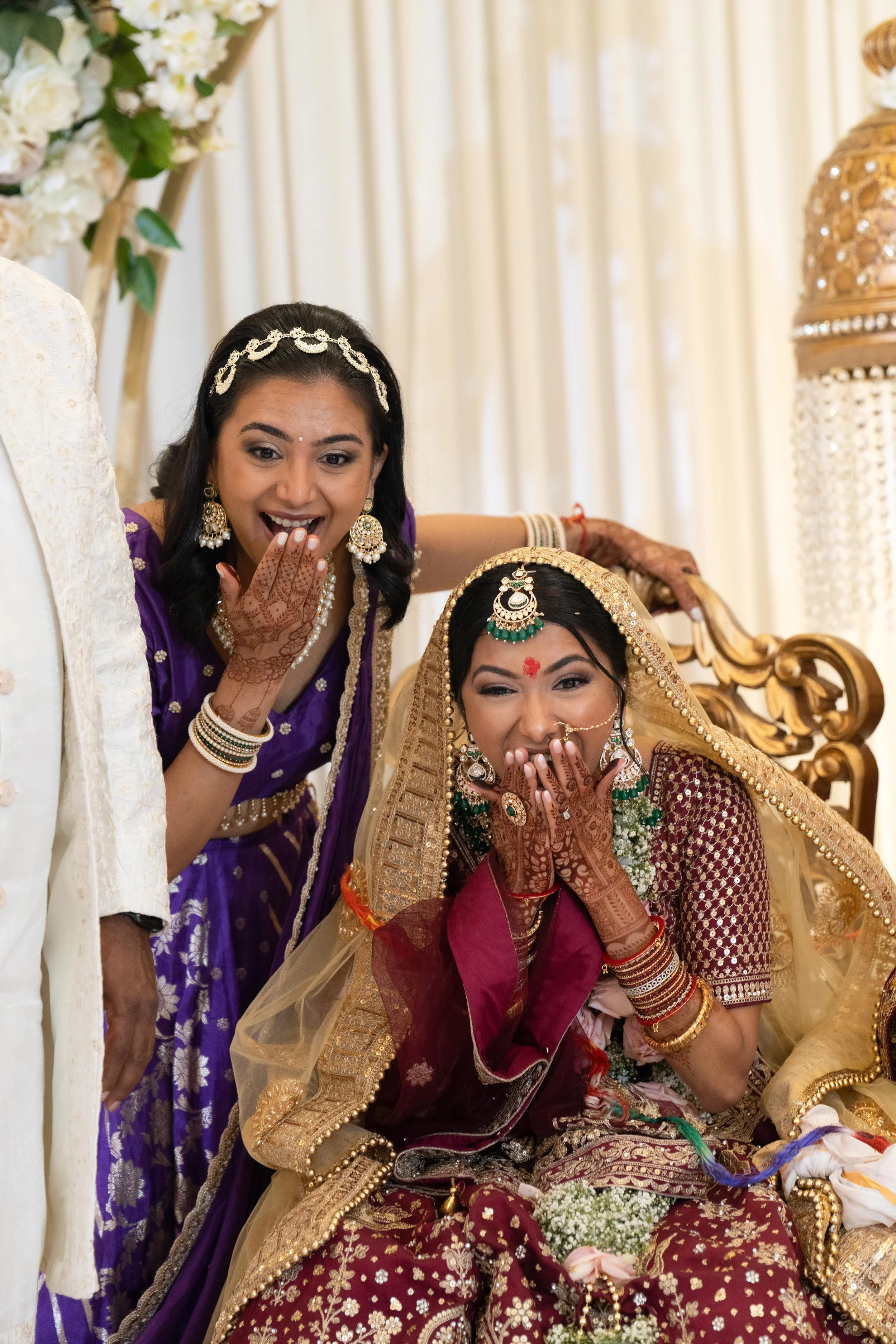 Indian bride in traditional attire with jewelry, seated on a decorated chair, with woman beside her making a surprised and joyful gesture.