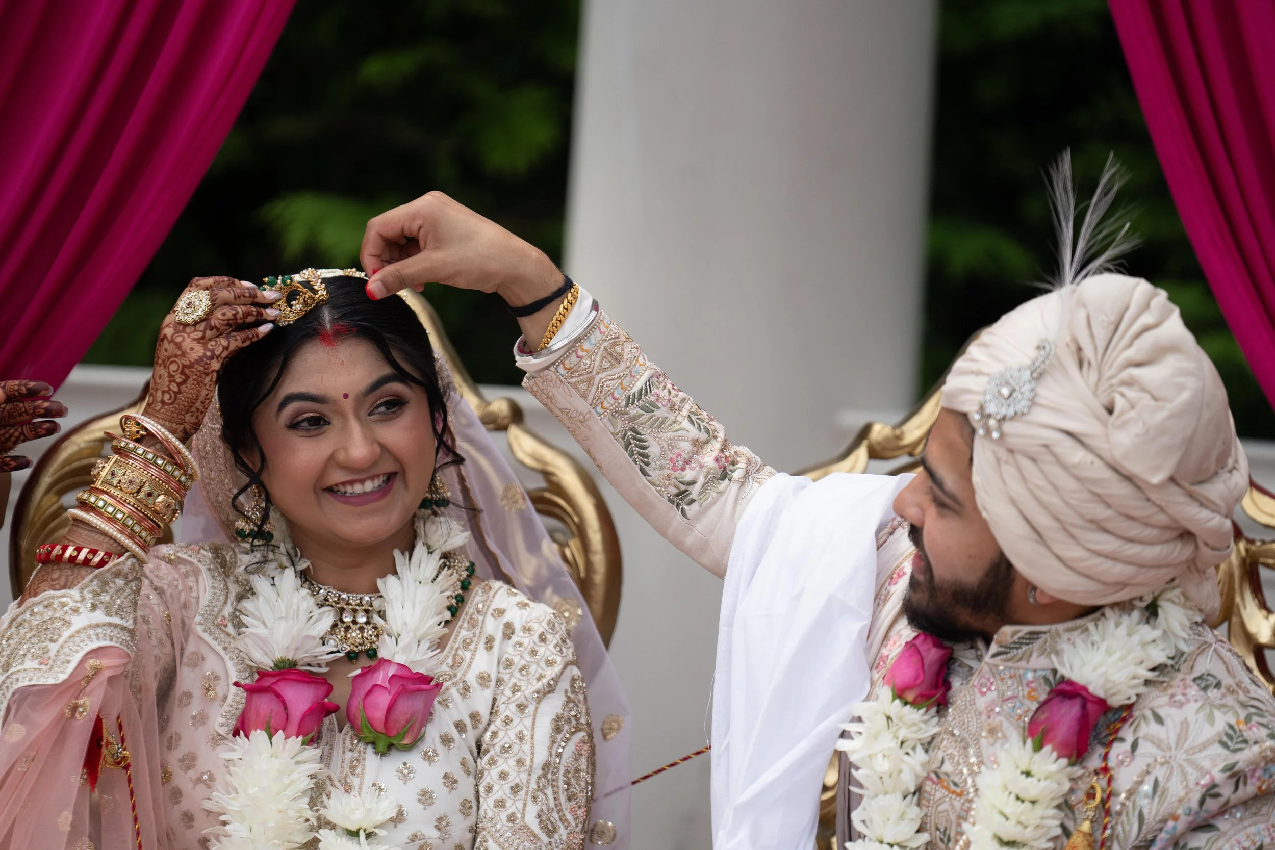 Indian bride smiling as groom places a necklace on her during wedding ceremony, with traditional attire and floral garlands.