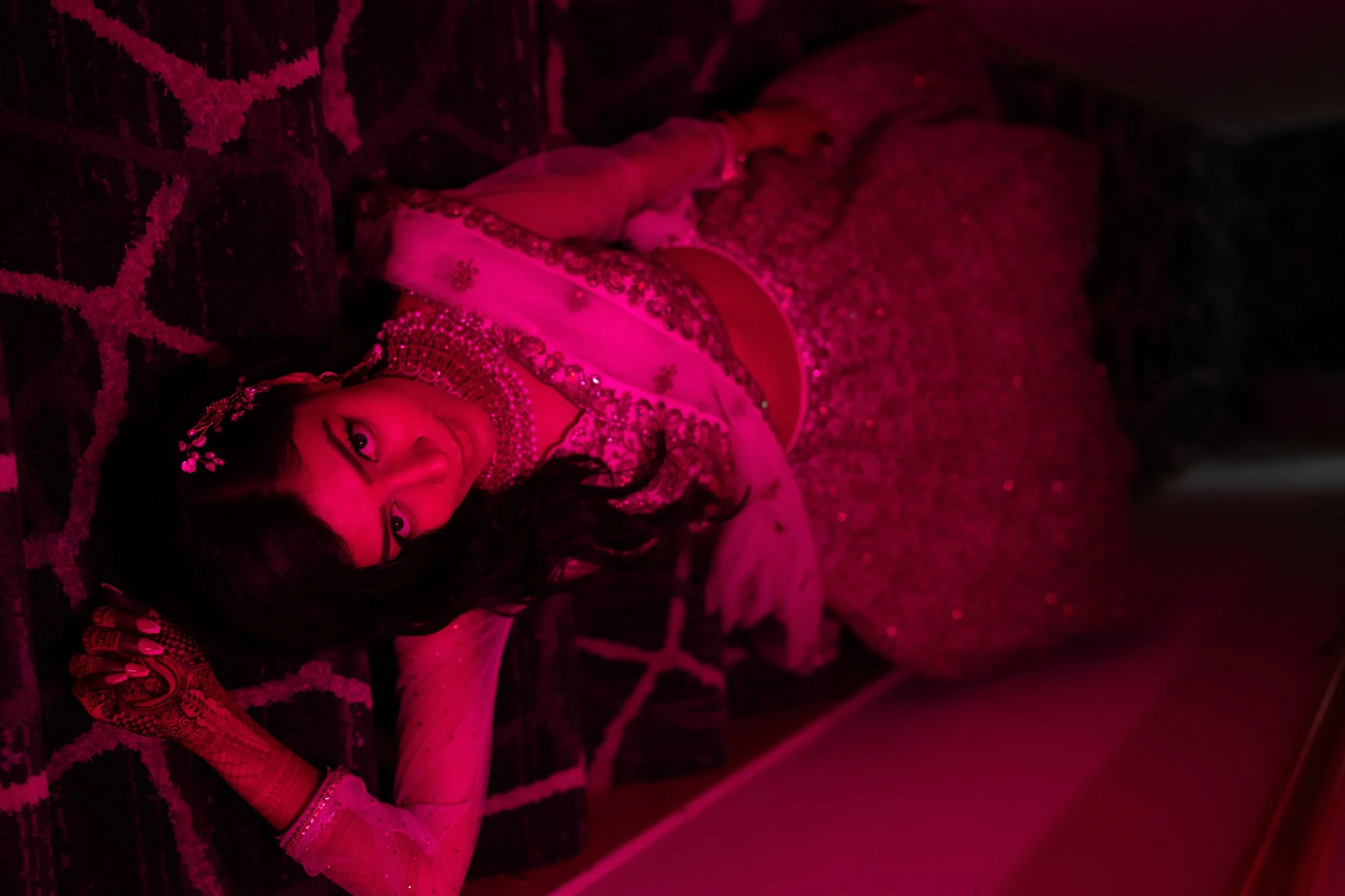 A Bride dressed in traditional, embellished Indian attire lying on a patterned carpet in a room with pink lighting, looking at the camera.