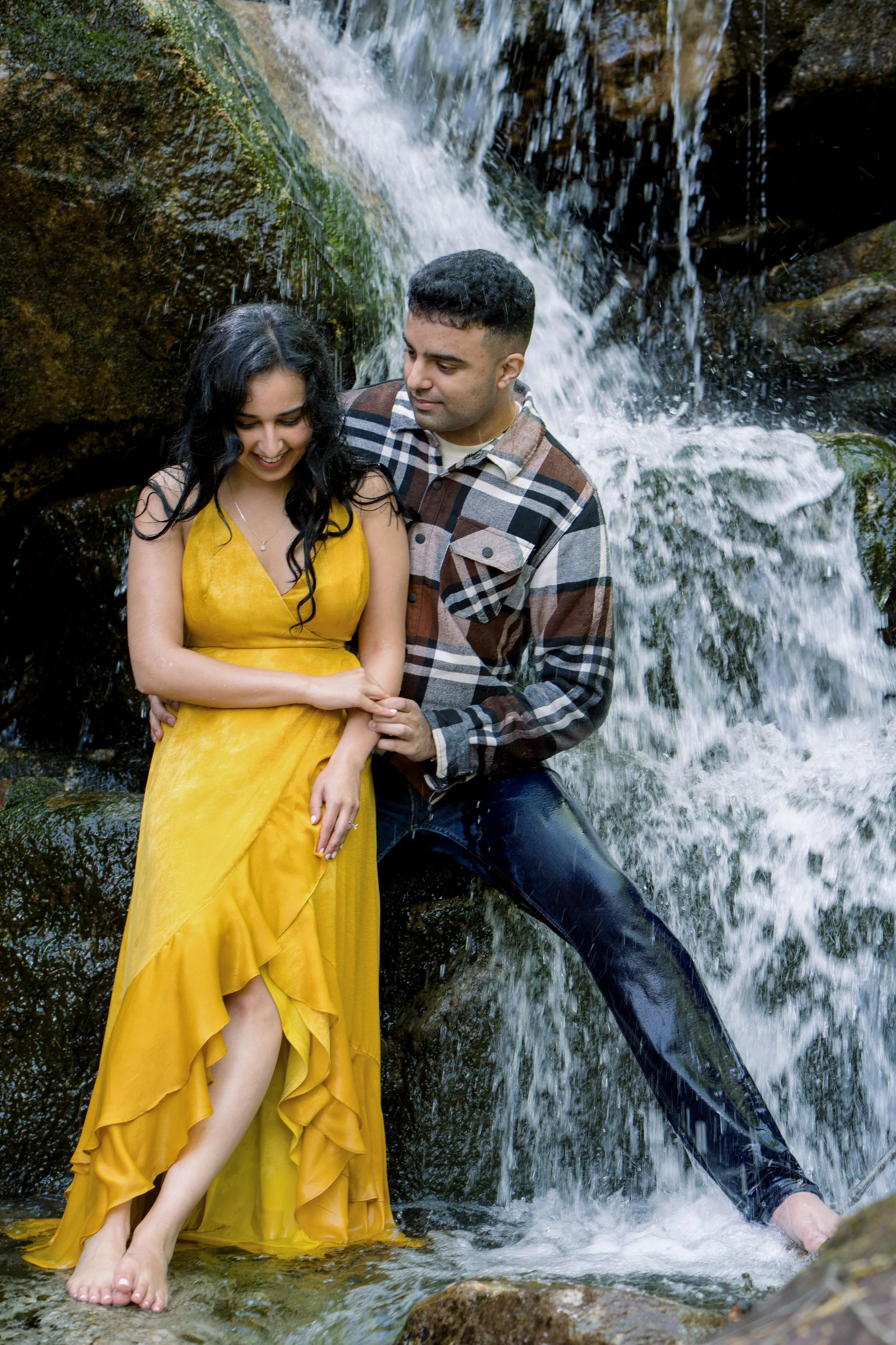 A man and woman standing in front of a small waterfall, with the man helping the woman stand on a rock while they hold hands and smile.