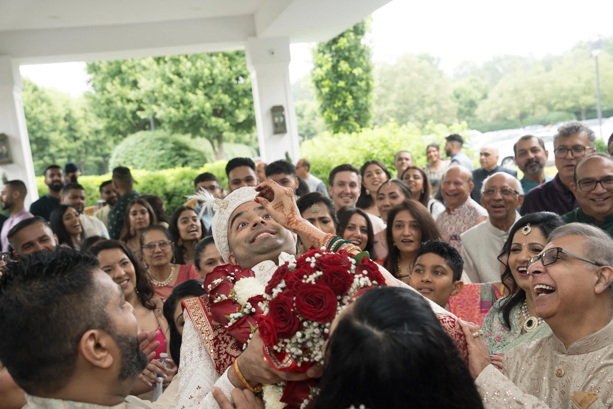 Indian groom in traditional attire with flower garland, smiling, surrounded by cheerful wedding guests at an outdoor celebration.