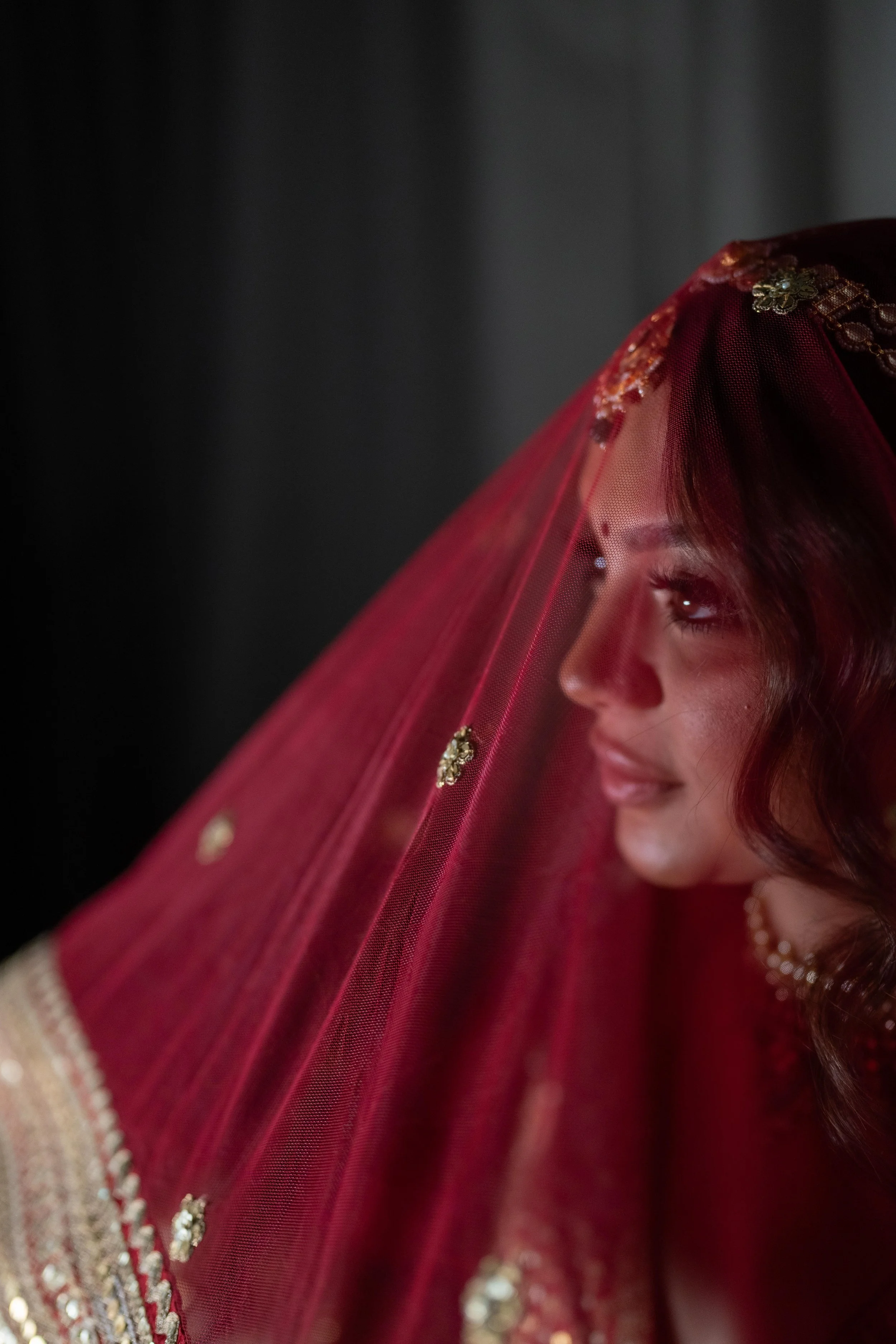 Close-up of a bride wearing a red bridal veil with gold embroidery, smiling, with her face turned sideways and dark background.