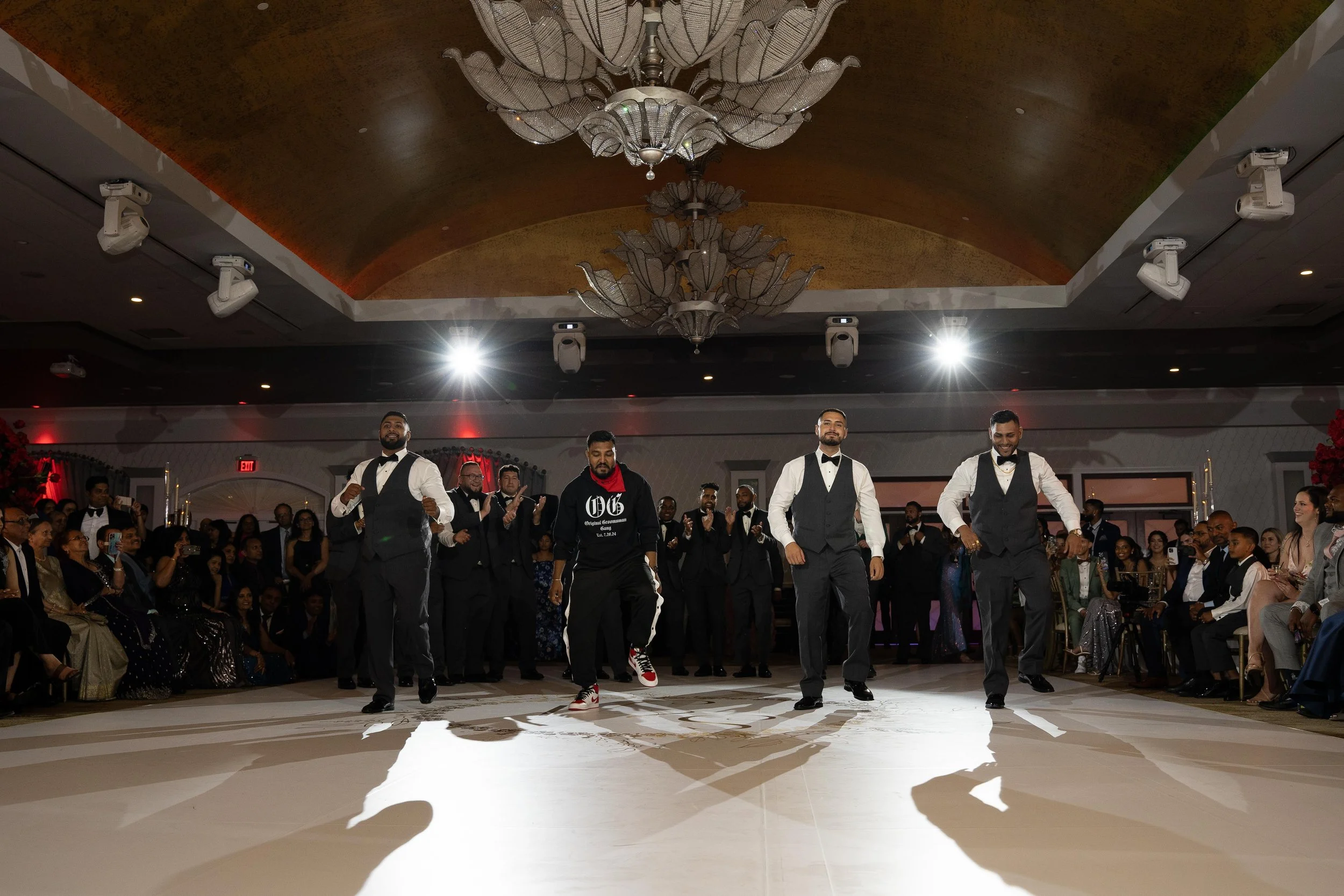 A groomsman dancing in tuxedos on a dance floor at a wedding or formal event, with a large audience seated around the floor and clapping.