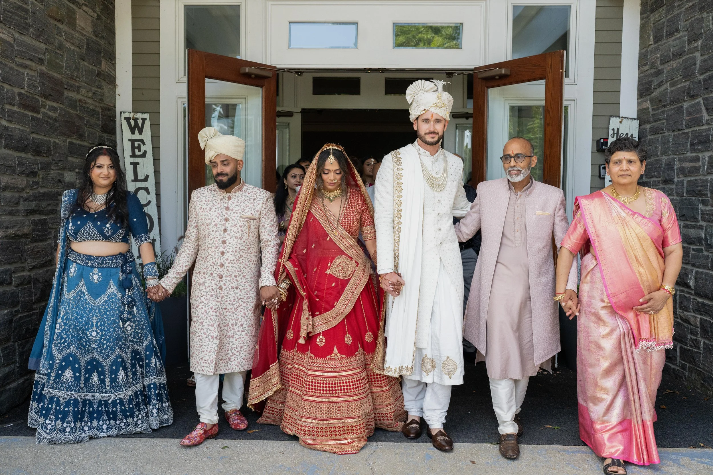 Indian wedding couple in traditional attire walking out of a building holding hands, surrounded by family members dressed in colorful, traditional Indian clothing.