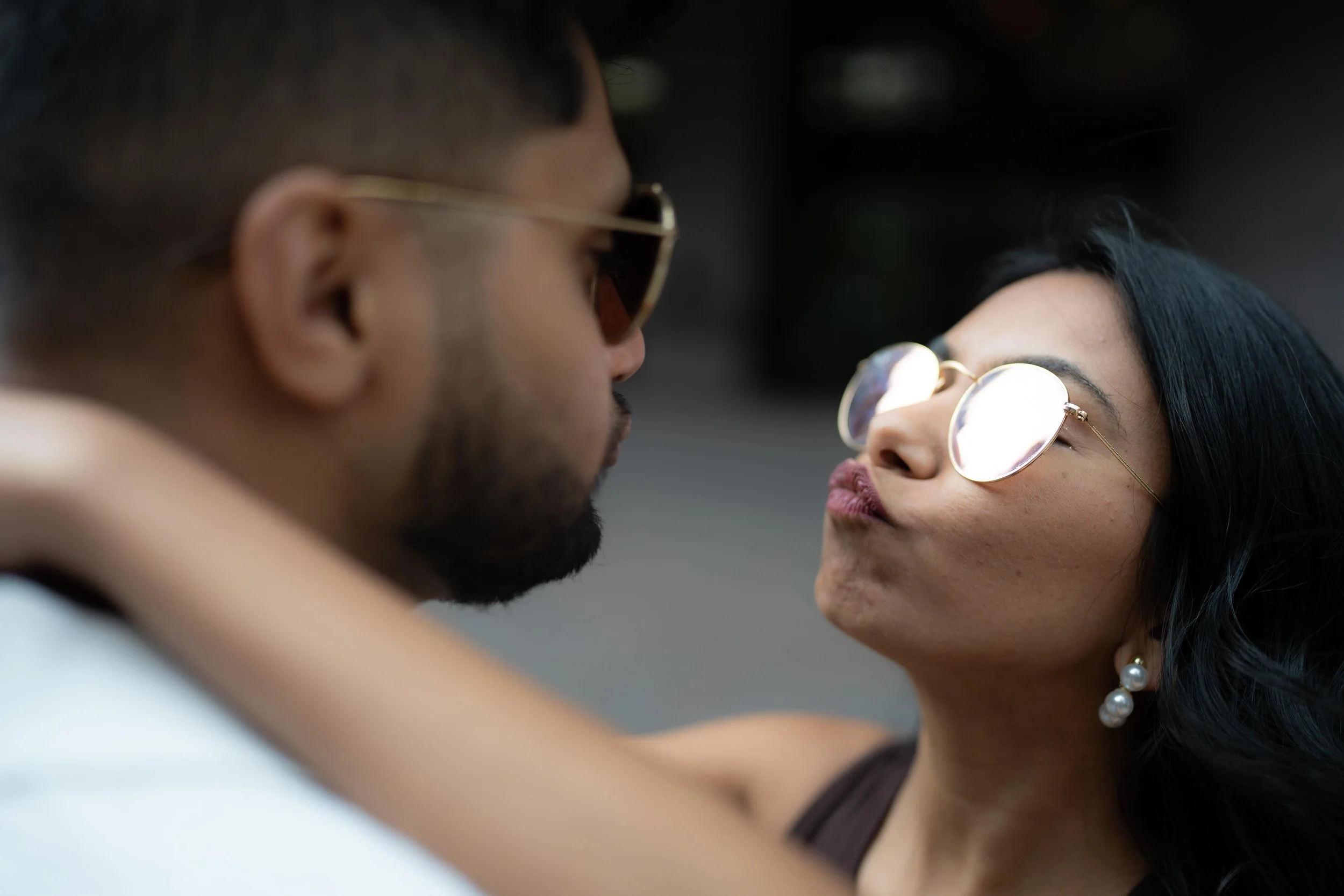 A groom and bride wearing sunglasses face each other closely, with the woman making a kissing face towards the man.