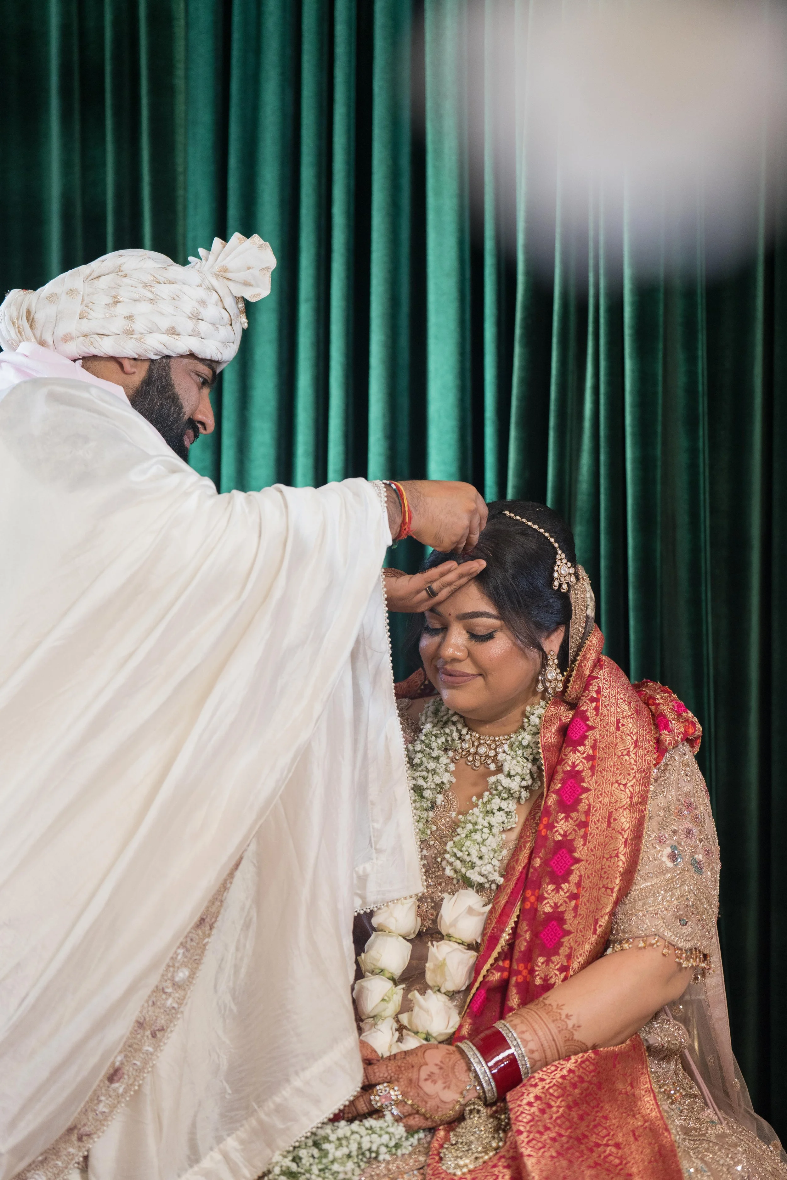 A groom performing a traditional Indian wedding ceremony on a bride, applying a mark on her forehead, with green curtains in the background.