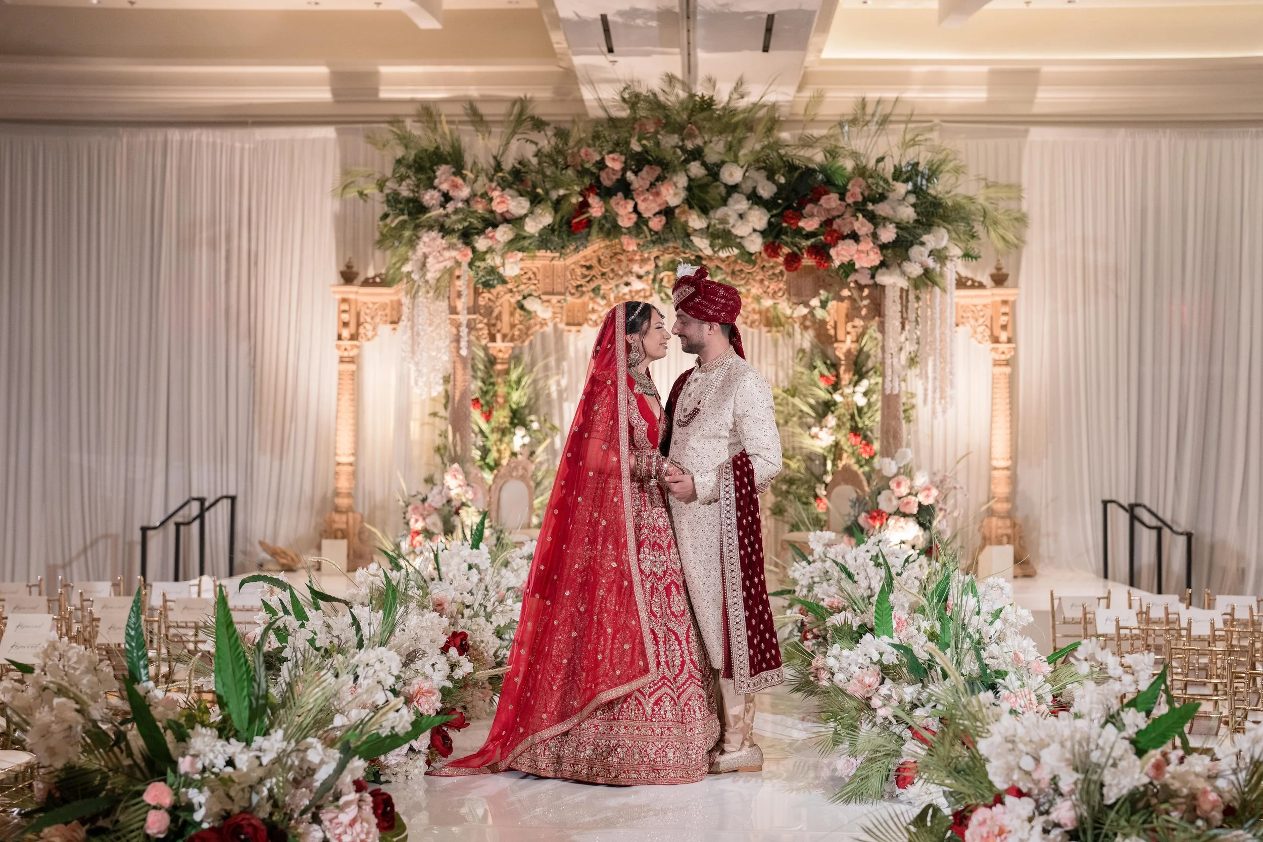 A bride and groom in traditional Indian wedding attire standing close, holding hands, and facing each other under a floral arch decorated with pink, white, and red flowers with greenery, during their wedding ceremony.