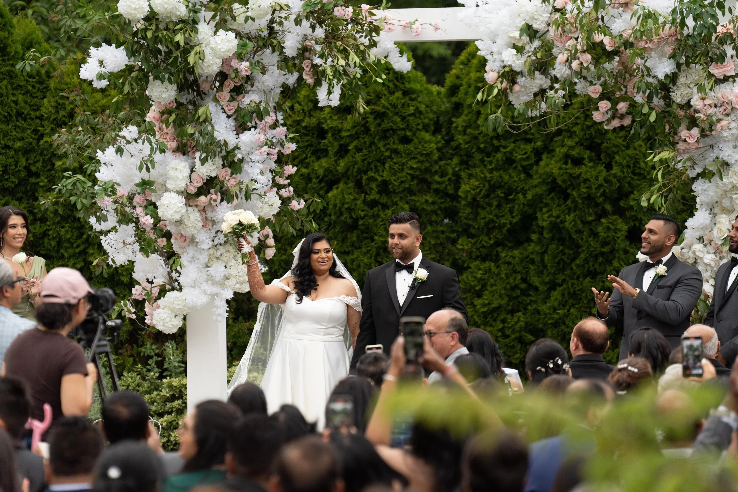 A wedding ceremony outdoors with the bride and groom under a floral arch. The bride is in a white wedding dress, holding a bouquet, and the groom is in a black tuxedo. Guests are seated and taking photos, with some clapping. The background features g