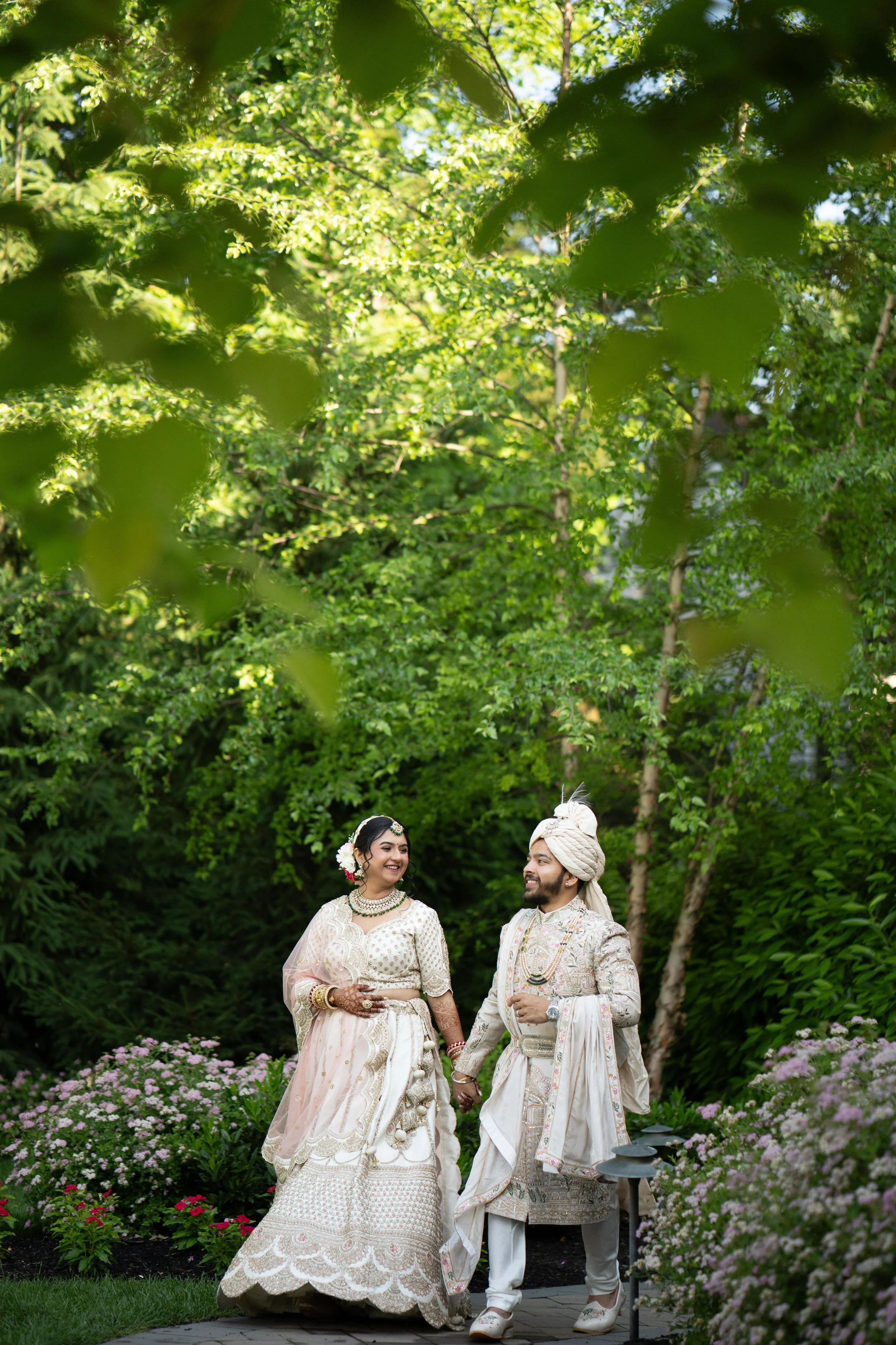 A newlywed couple in traditional Indian wedding attire holding hands and smiling at each other in a lush garden.