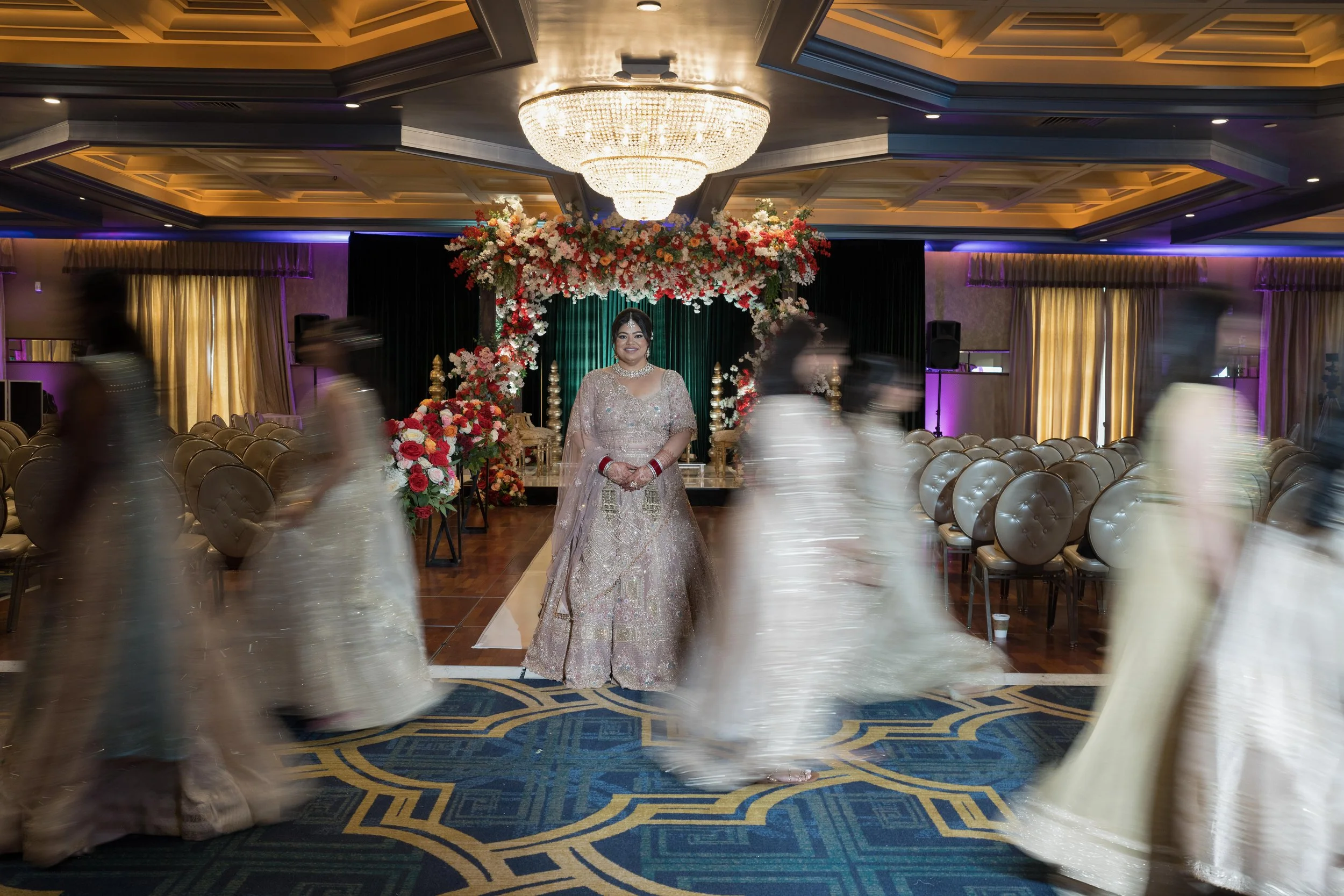 A bride in traditional Indian dress stands in the center of a decorated wedding hall surrounded by blurred dancers in white and gold outfits.