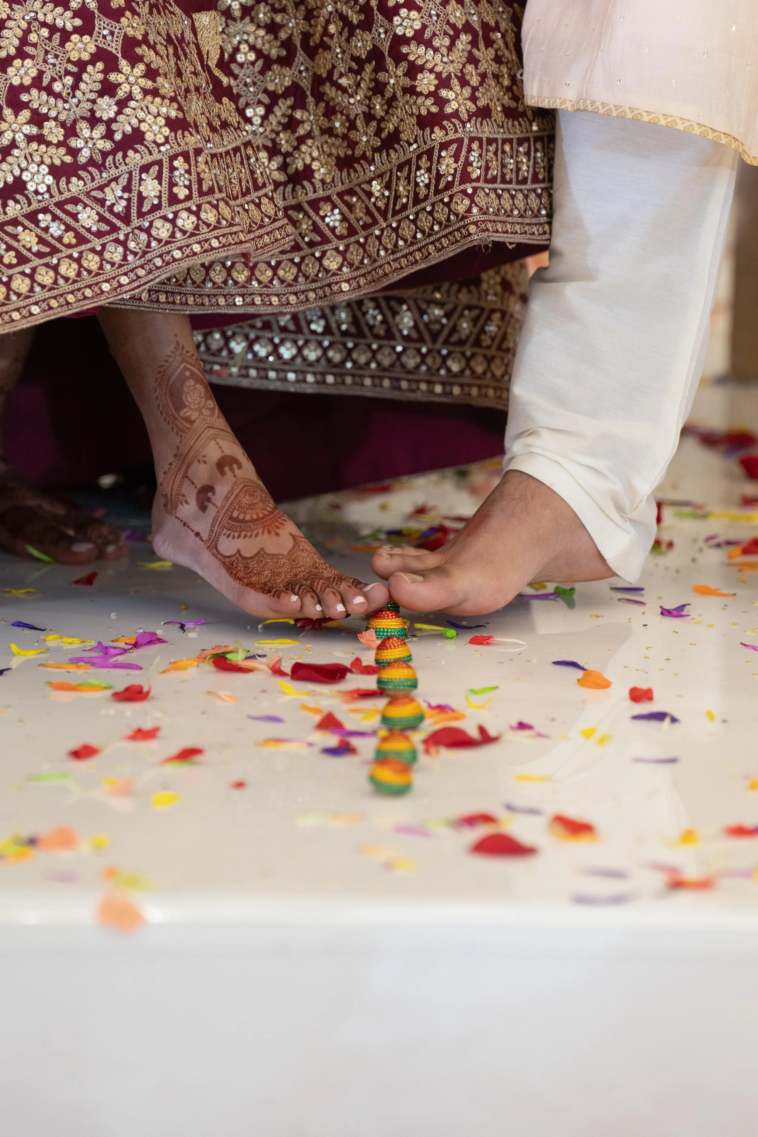 Close-up of a traditional Indian wedding ceremony showing a bride with henna on her hand and a groom in white, both touching a decorative, beaded line on the floor amid colorful flower petals.