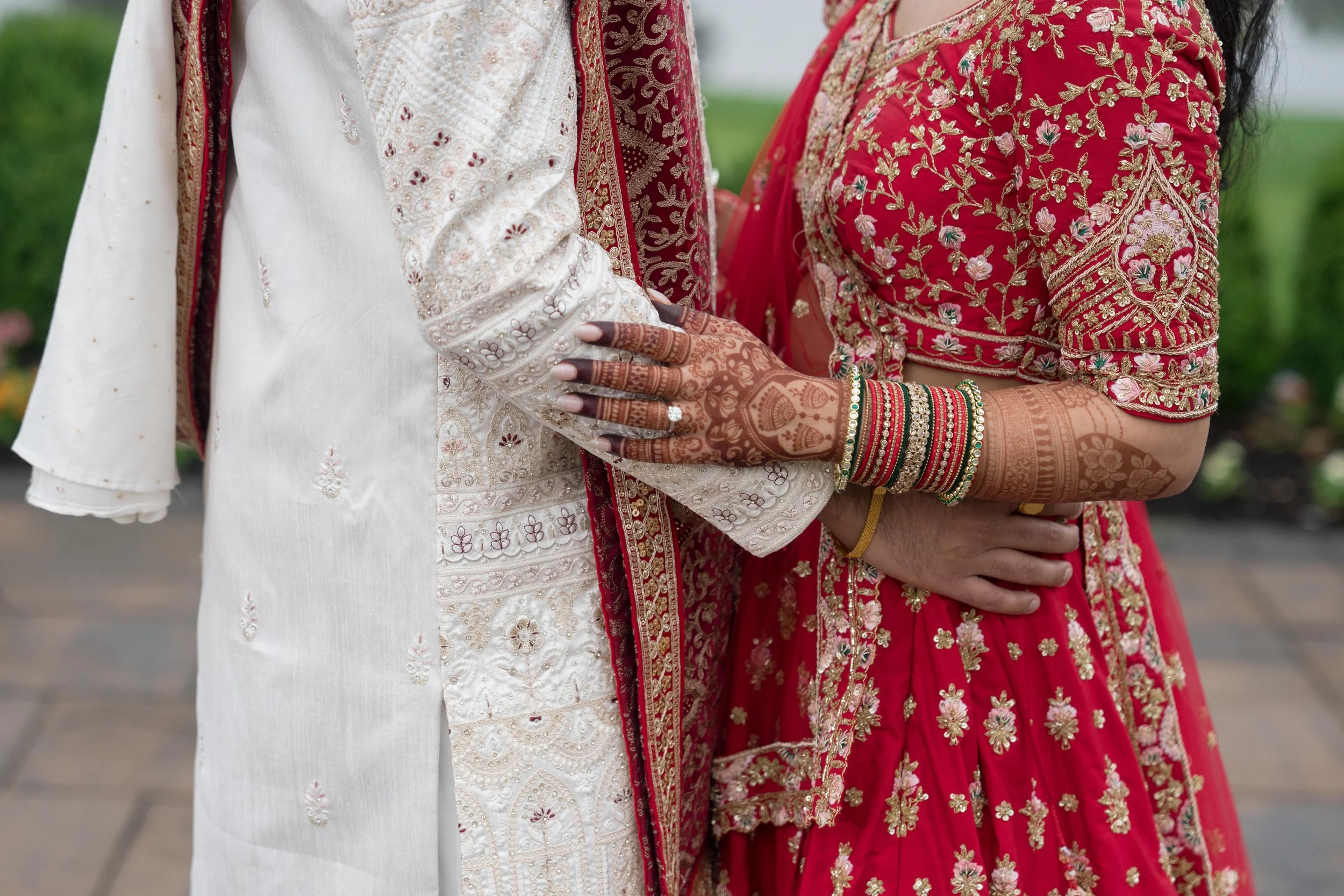 Close-up of a bride and groom holding hands during a wedding, with the bride wearing traditional red and gold embroidered attire, bangles, and henna, and the groom in a white embroidered outfit.