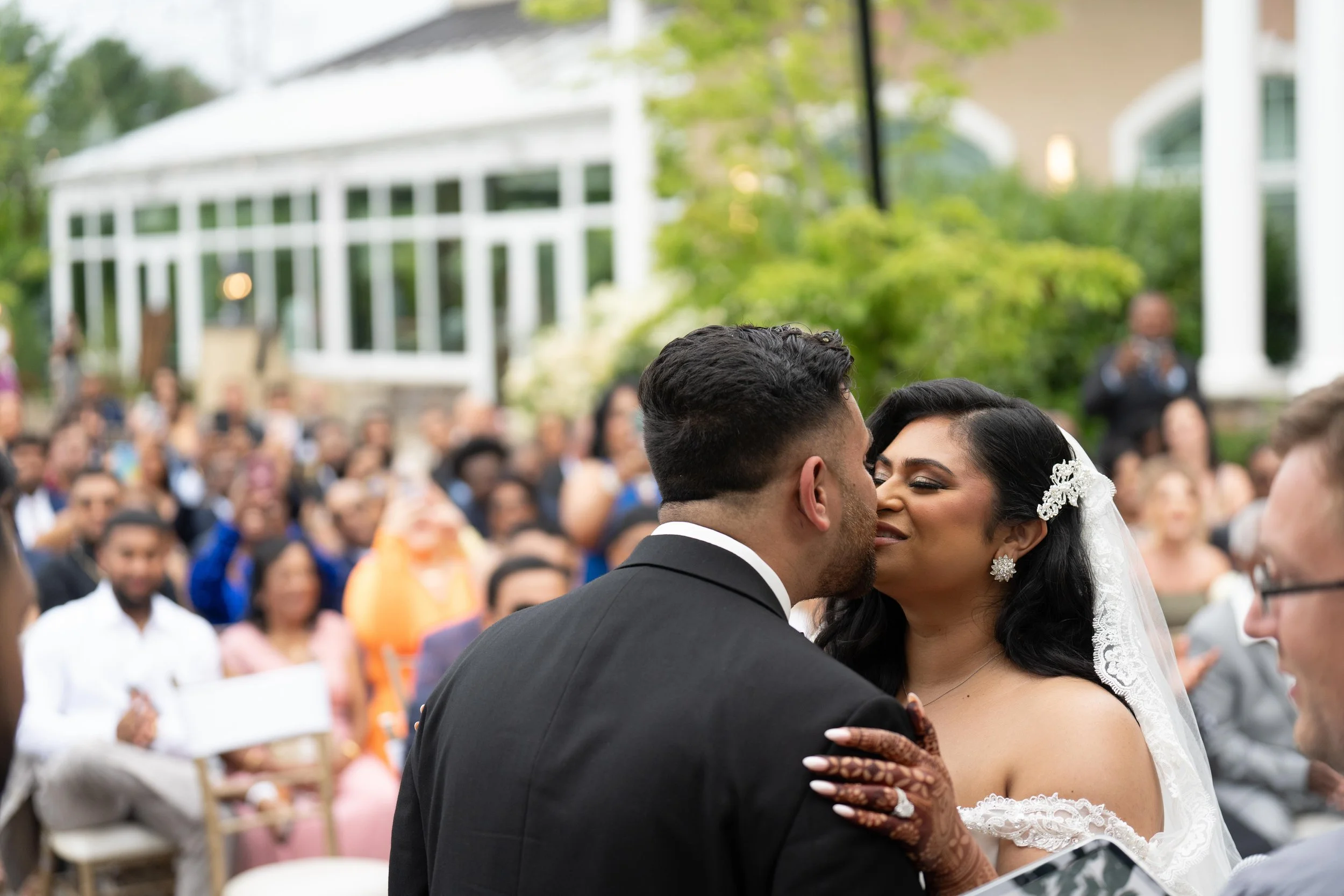 A couple getting married, sharing a kiss during their outdoor wedding ceremony, with a crowd of guests in the background.
