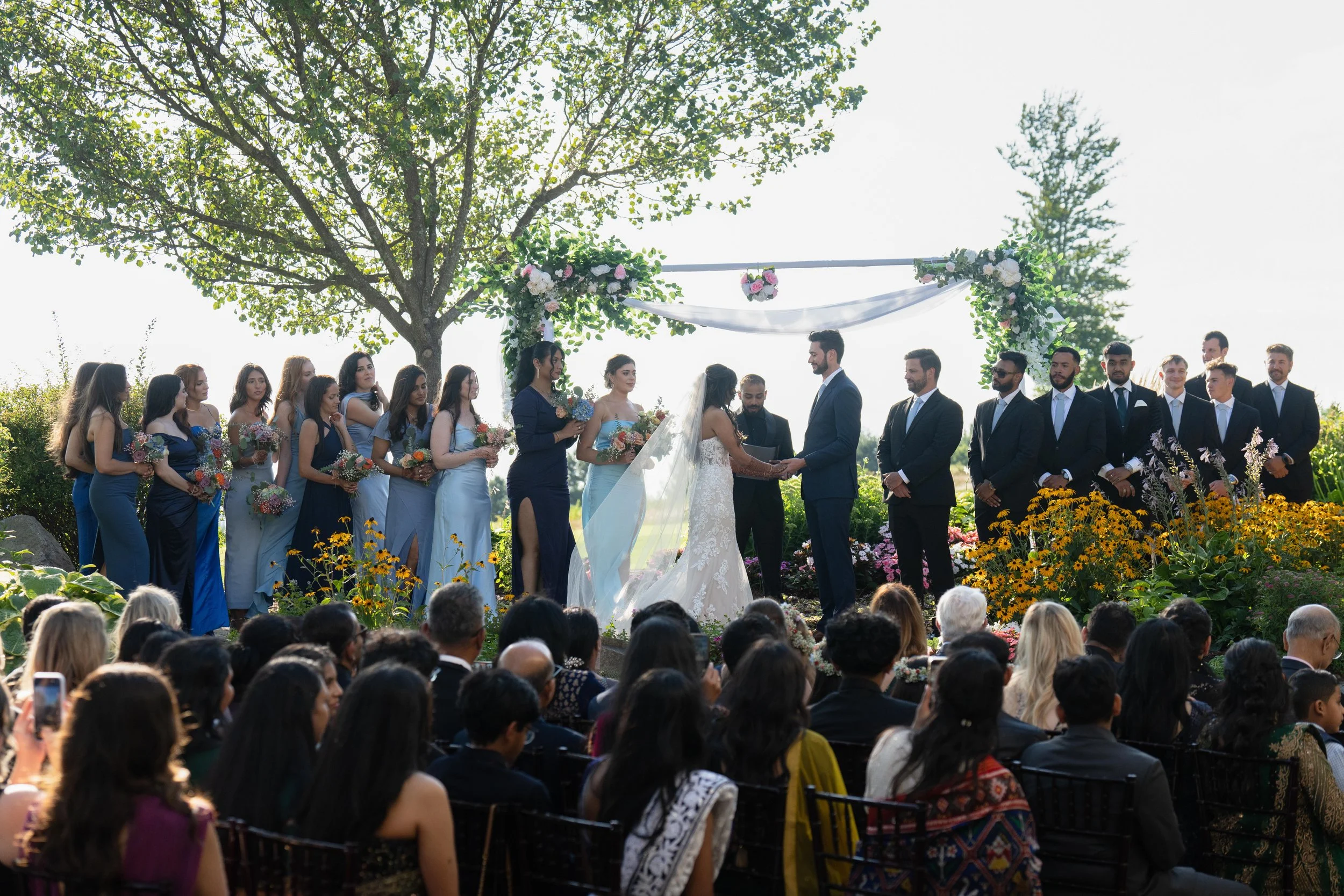 A wedding ceremony taking place outdoors under a large tree with sunlight filtering through. The bride and groom are exchanging vows, surrounded by bridesmaids and groomsmen, with an audience seated in front watching.