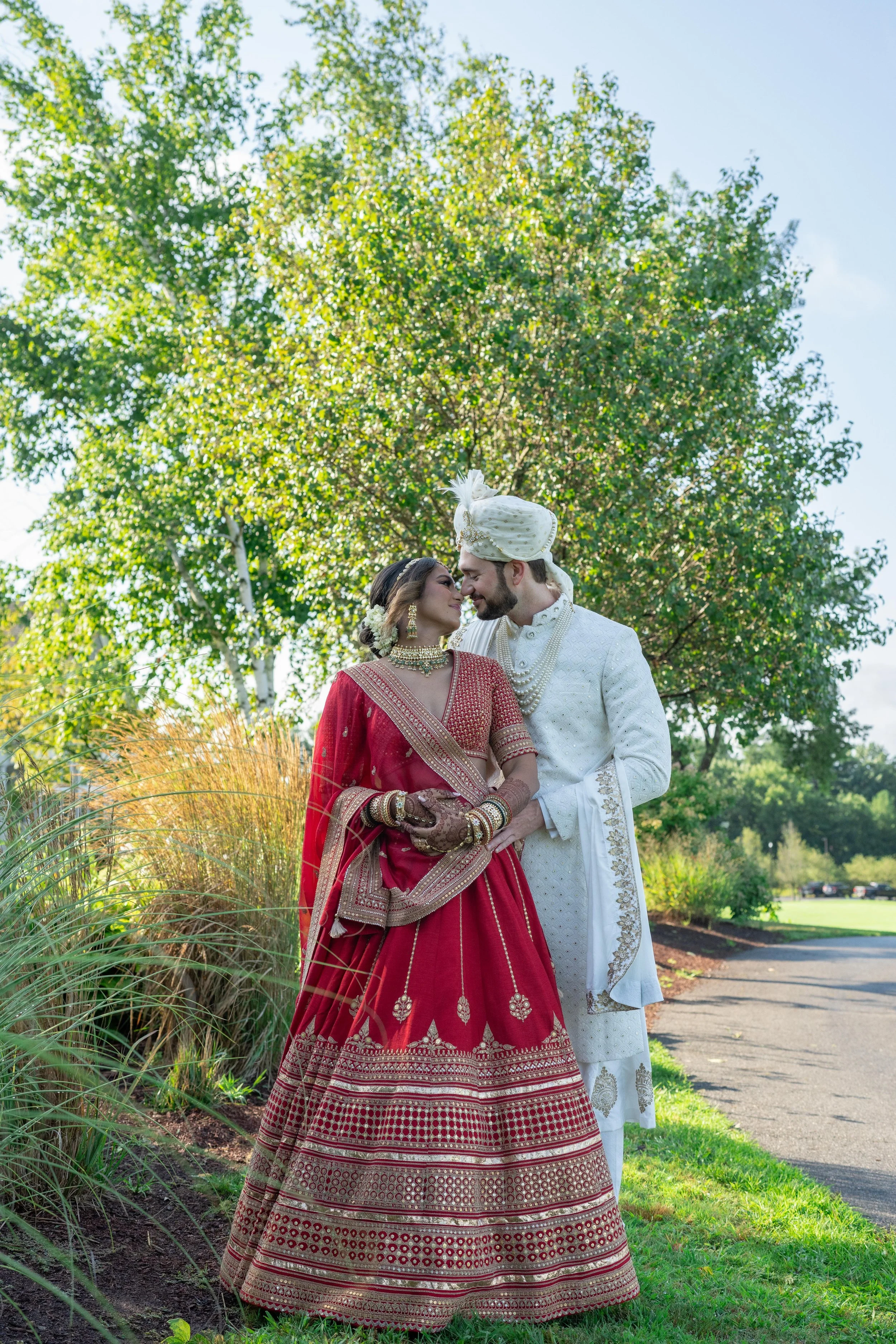 A couple dressed in traditional Indian wedding attire standing outdoors, touching foreheads, with green trees and a blue sky in the background.
