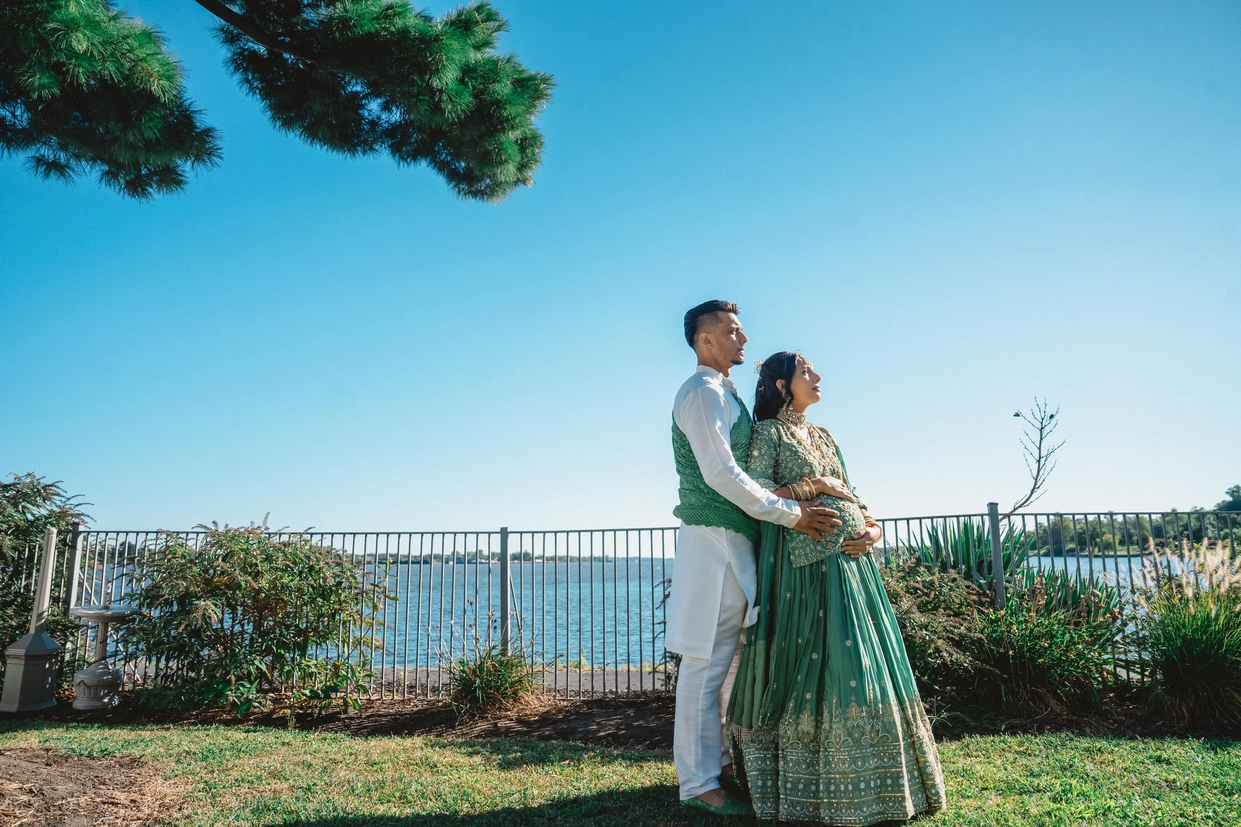 A couple dressed in traditional Indian attire standing outdoors by a waterfront, with the man gently holding the woman's pregnant belly, both gazing into the distance.