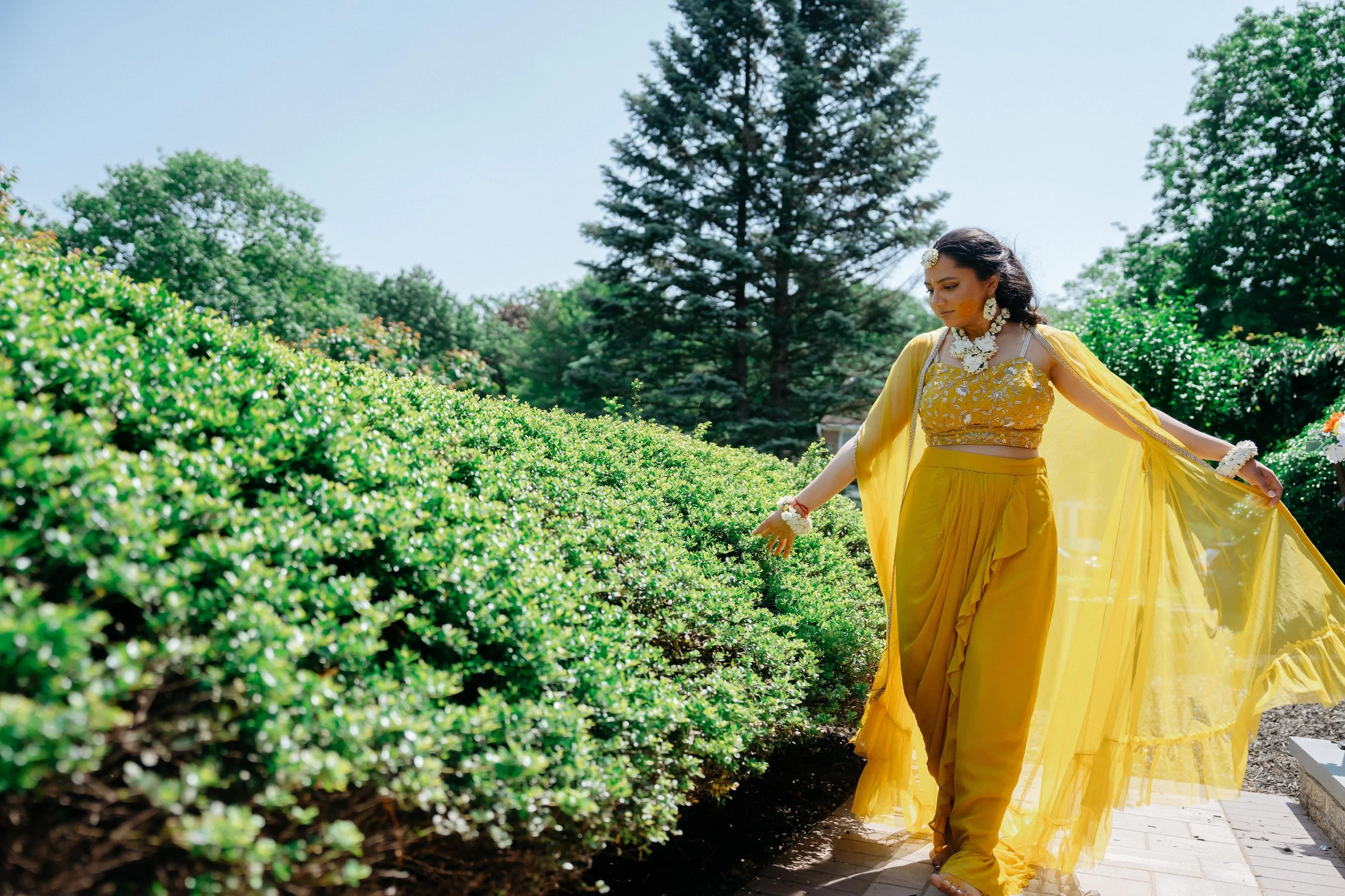 A bride in a yellow traditional Indian outfit standing outdoors near green bushes and trees