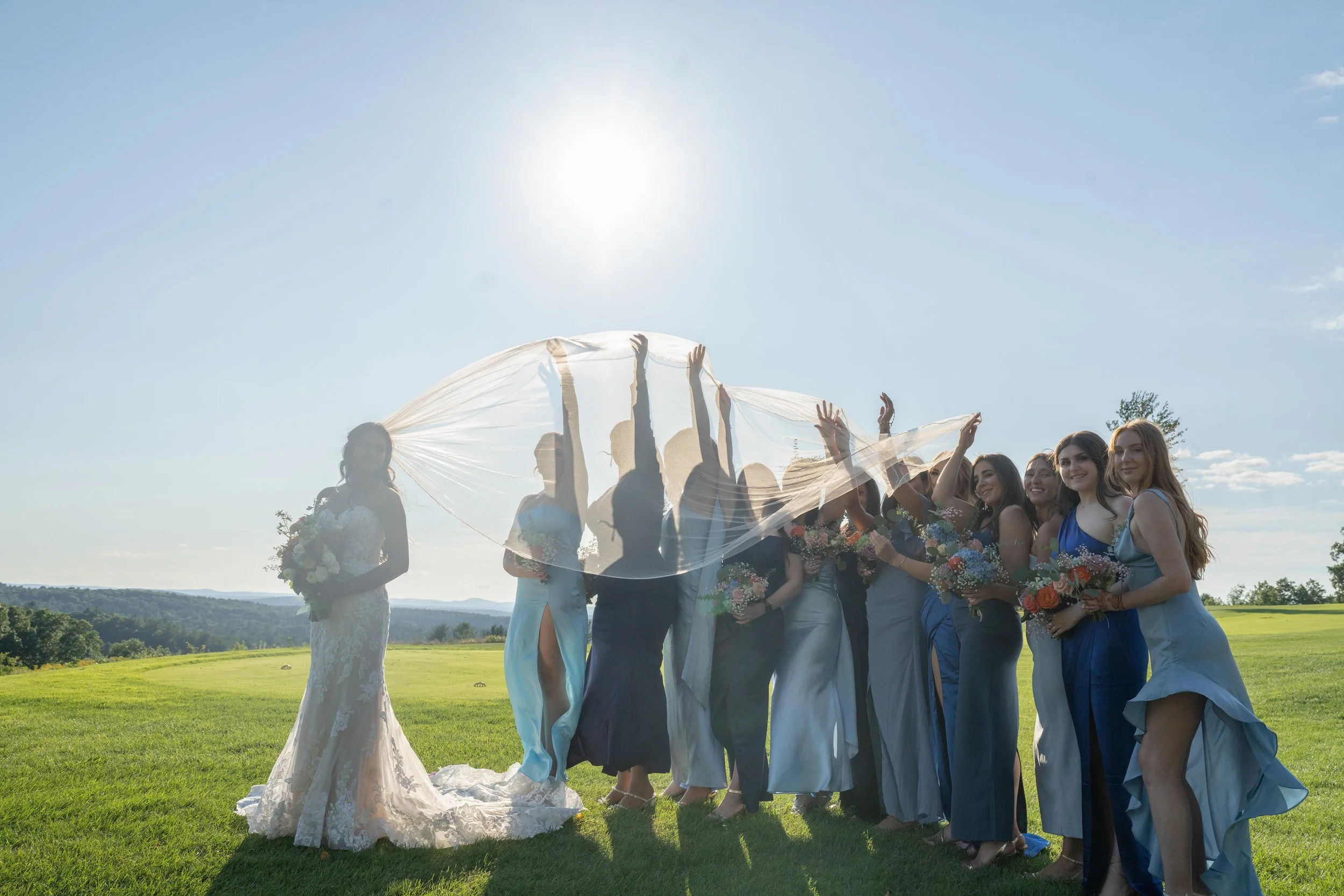 A bride in a lace wedding gown holding a bouquet of flowers standing on a grassy field, surrounded by bridesmaids in blue dresses holding bouquets, with some of them raising arms and a transparent veil in the sunlight.