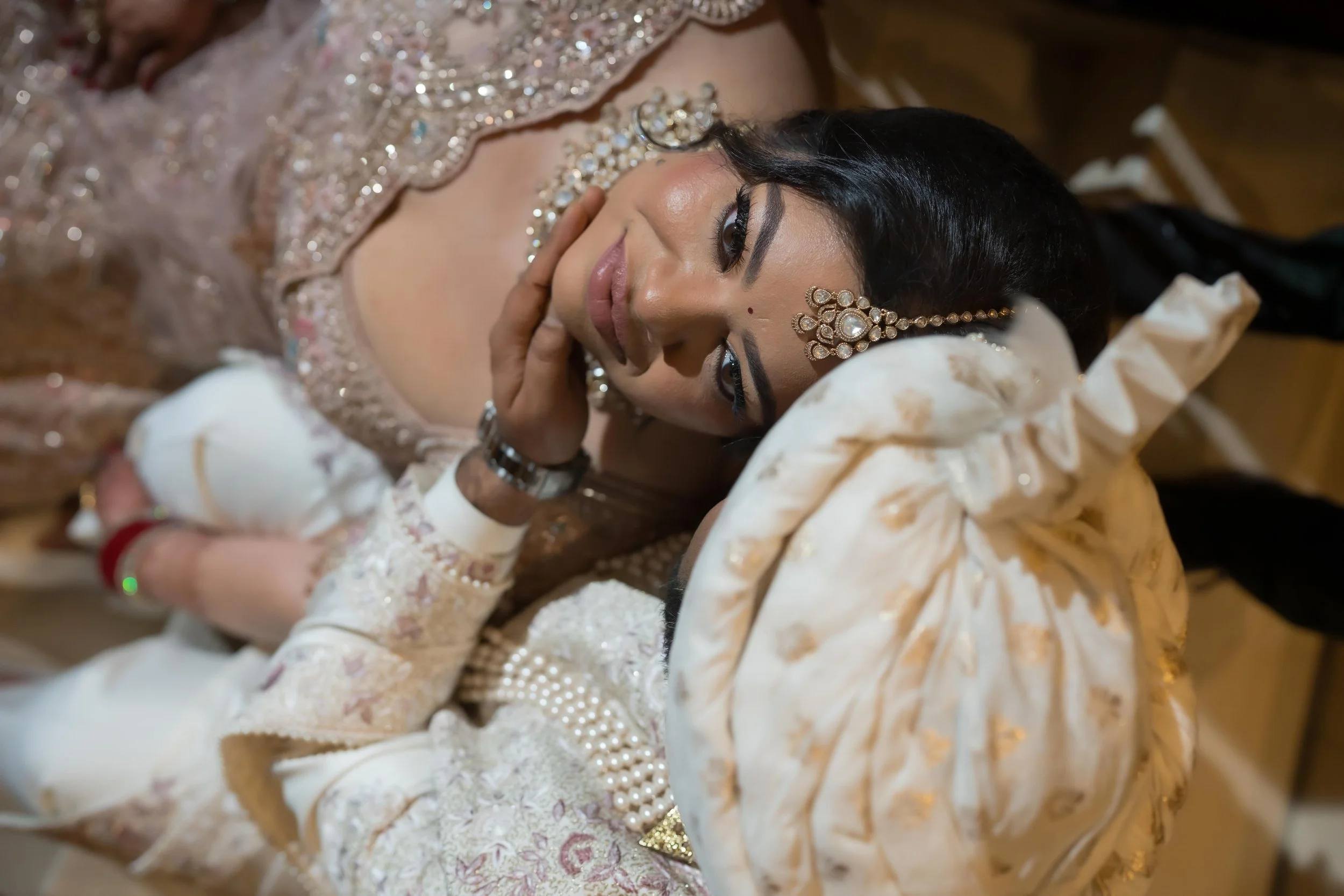 A bride lying down, dressed in traditional Indian wedding attire with jewelry, posing with an elegant expression.