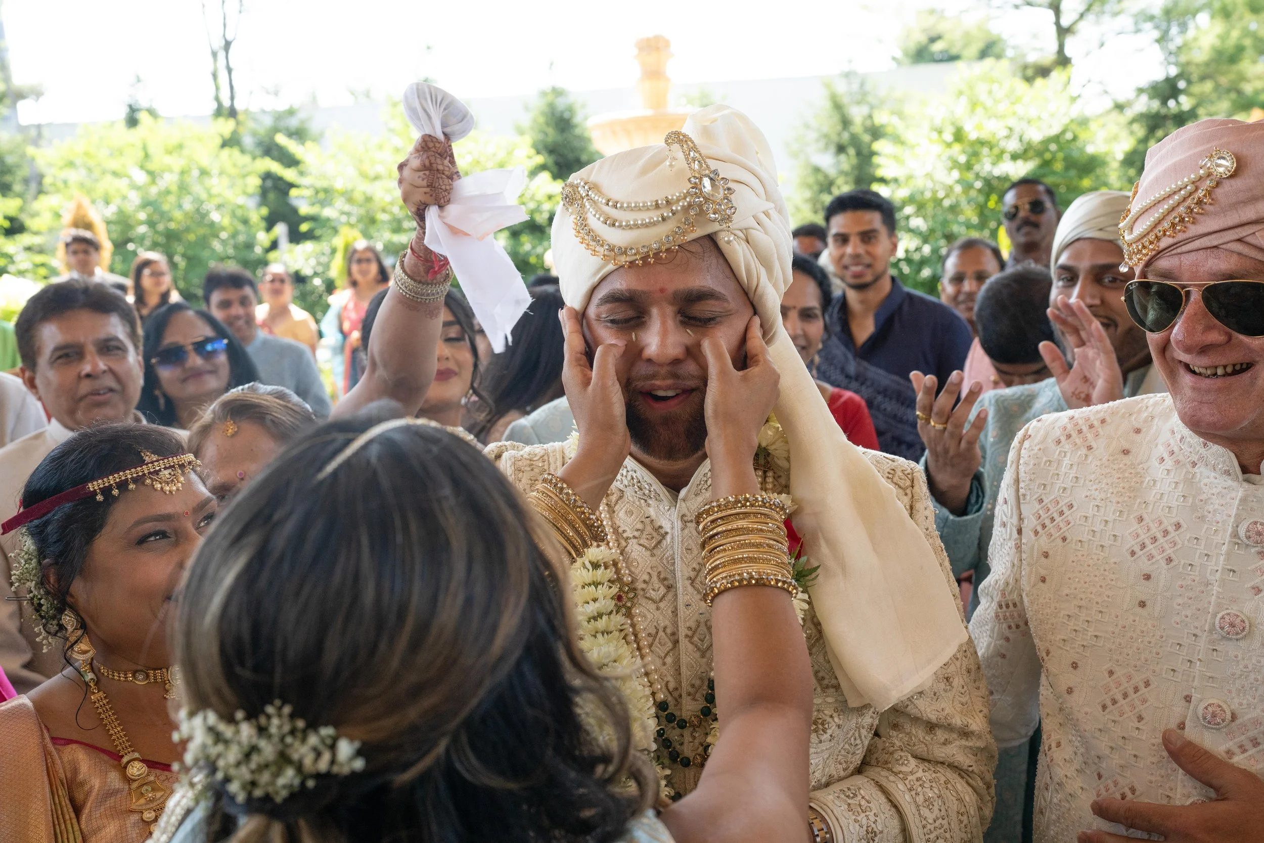 A traditional Indian wedding ceremony with the groom in ornate cream and gold attire, wearing a turban decorated with jewels, being blessed by the bride who is applying ceremonial marks on his face. The scene is lively with friends and family dressed