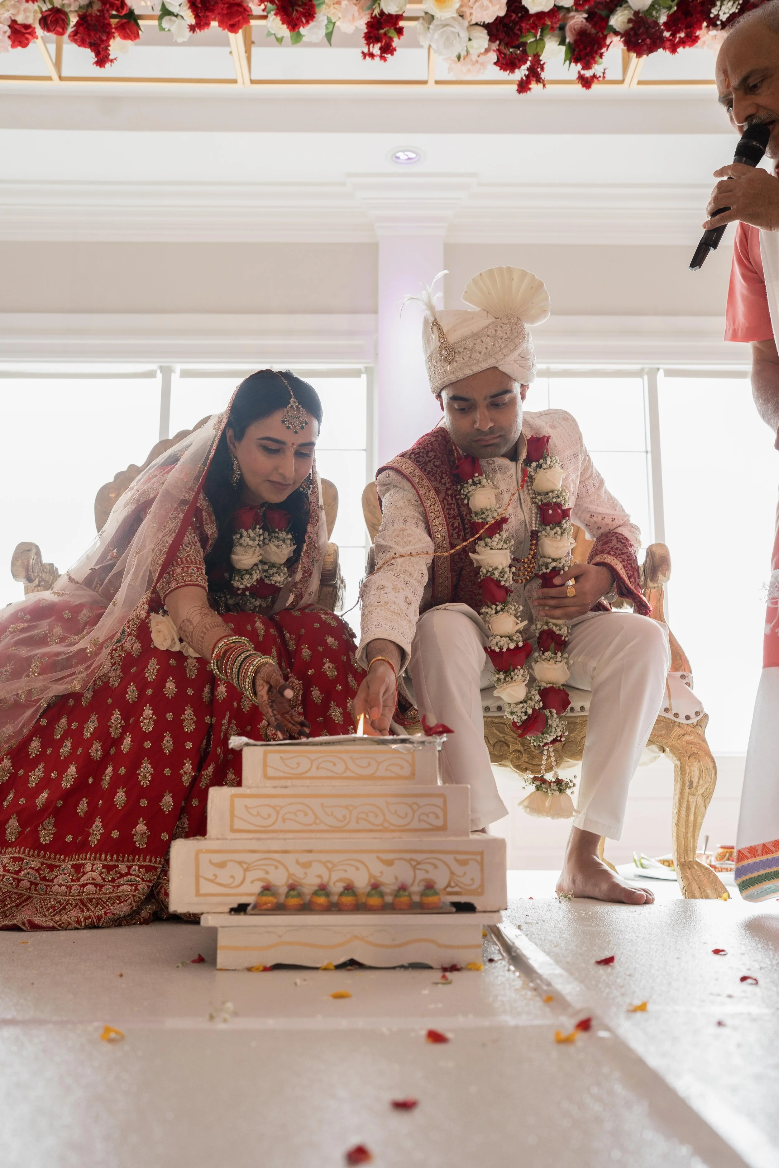 A bride and groom participating in a traditional Indian wedding ceremony, lighting a ceremonial fire with a priest present, all dressed in traditional attire with floral garlands.