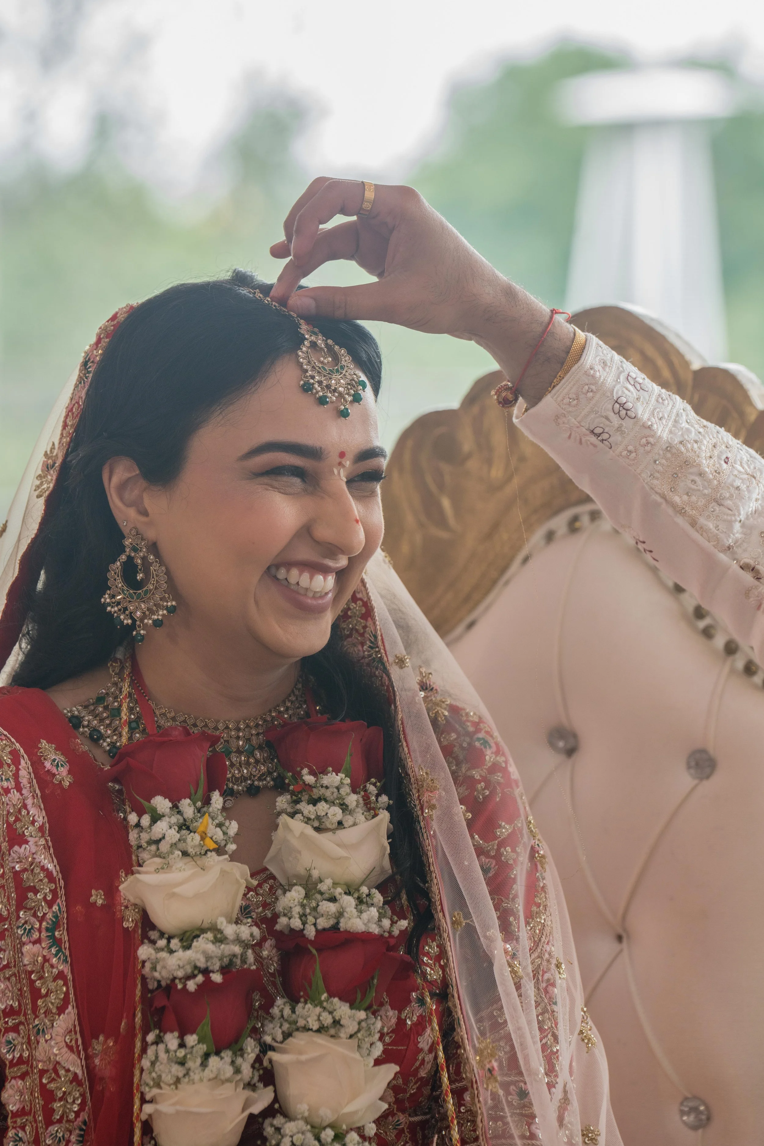 A bride dressed in traditional Indian wedding attire smiling as her forehead is being decorated by a man during a wedding ceremony.