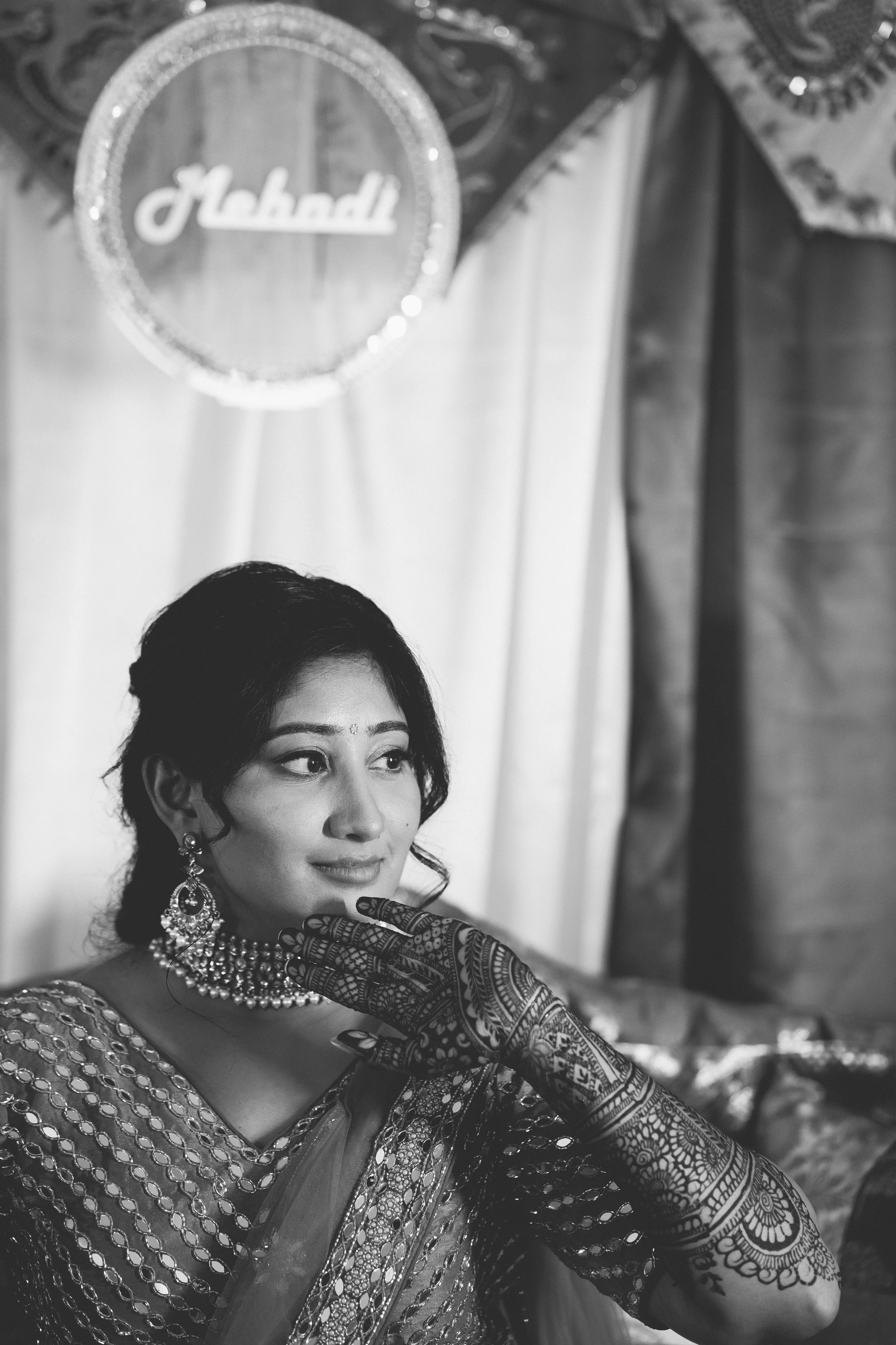 A Bride dressed in traditional Indian attire, wearing jewelry and henna designs on her hand, posing at a celebration or wedding, with a heart-shaped frame and the word 'Madh'.