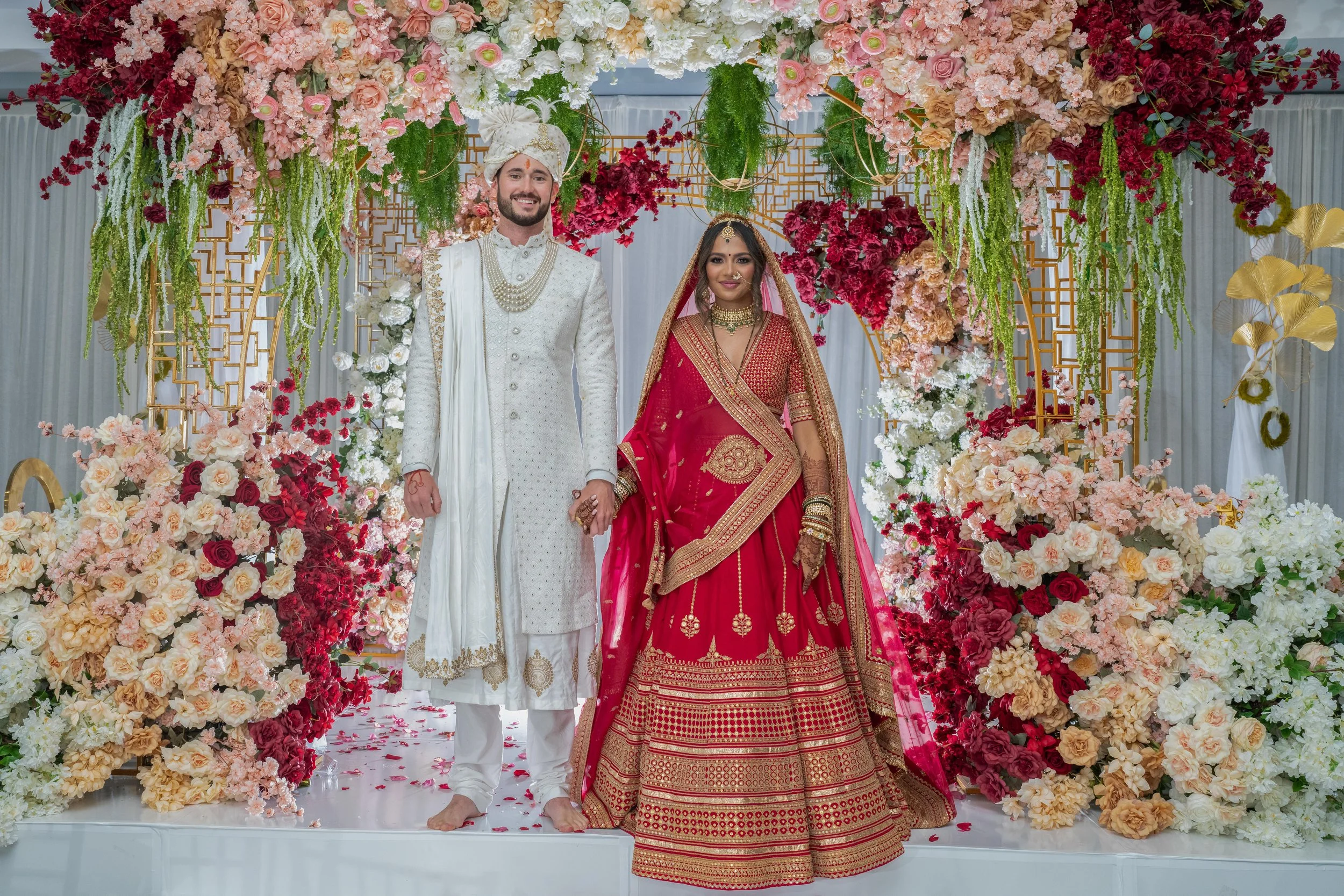A newlywed couple dressed in traditional Indian wedding attire stands hand in hand on a decorated wedding stage with an elaborate floral backdrop of pink, white, and red flowers.