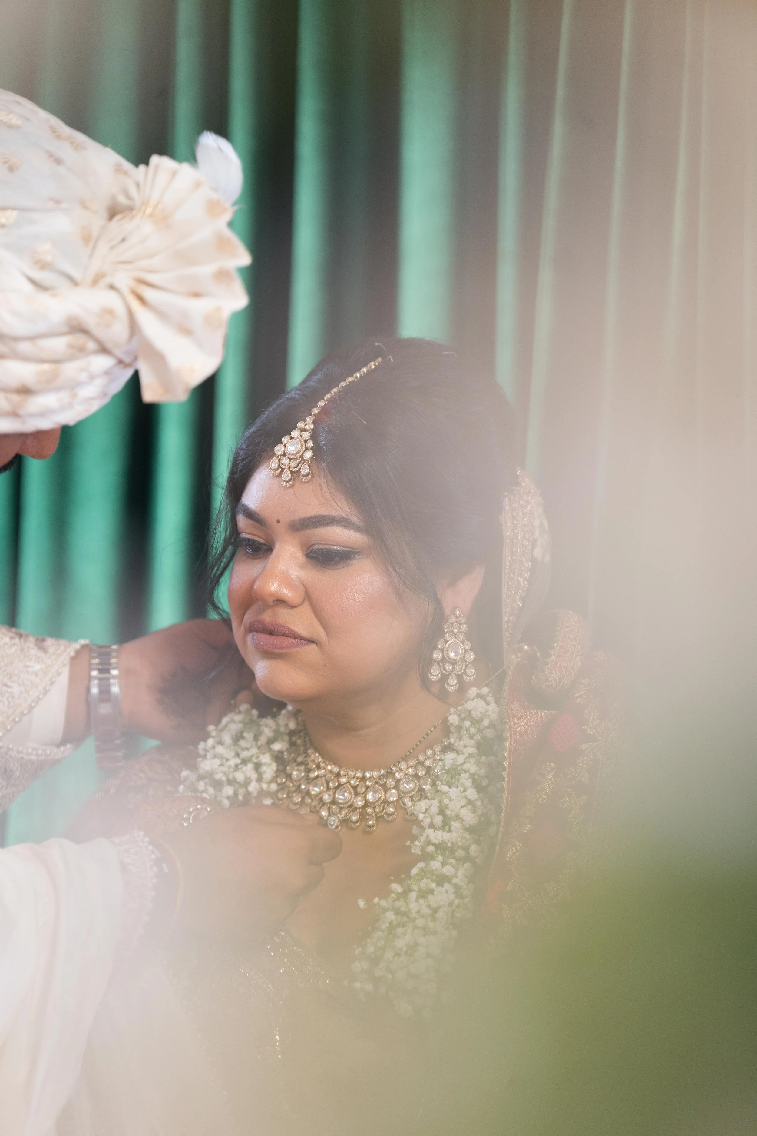A bride dressed in traditional Indian wedding attire, wearing jewelry and a garland of flowers, with a groom adjusting her necklace during a wedding ceremony