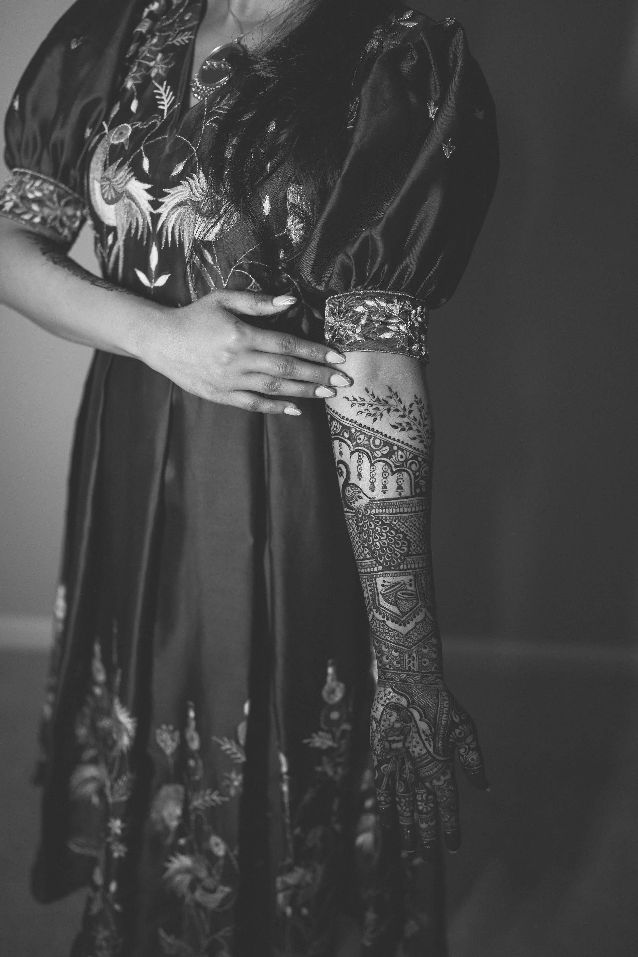 A woman wearing a dark, embroidered satin dress showing a detailed henna tattoo on her forearm.