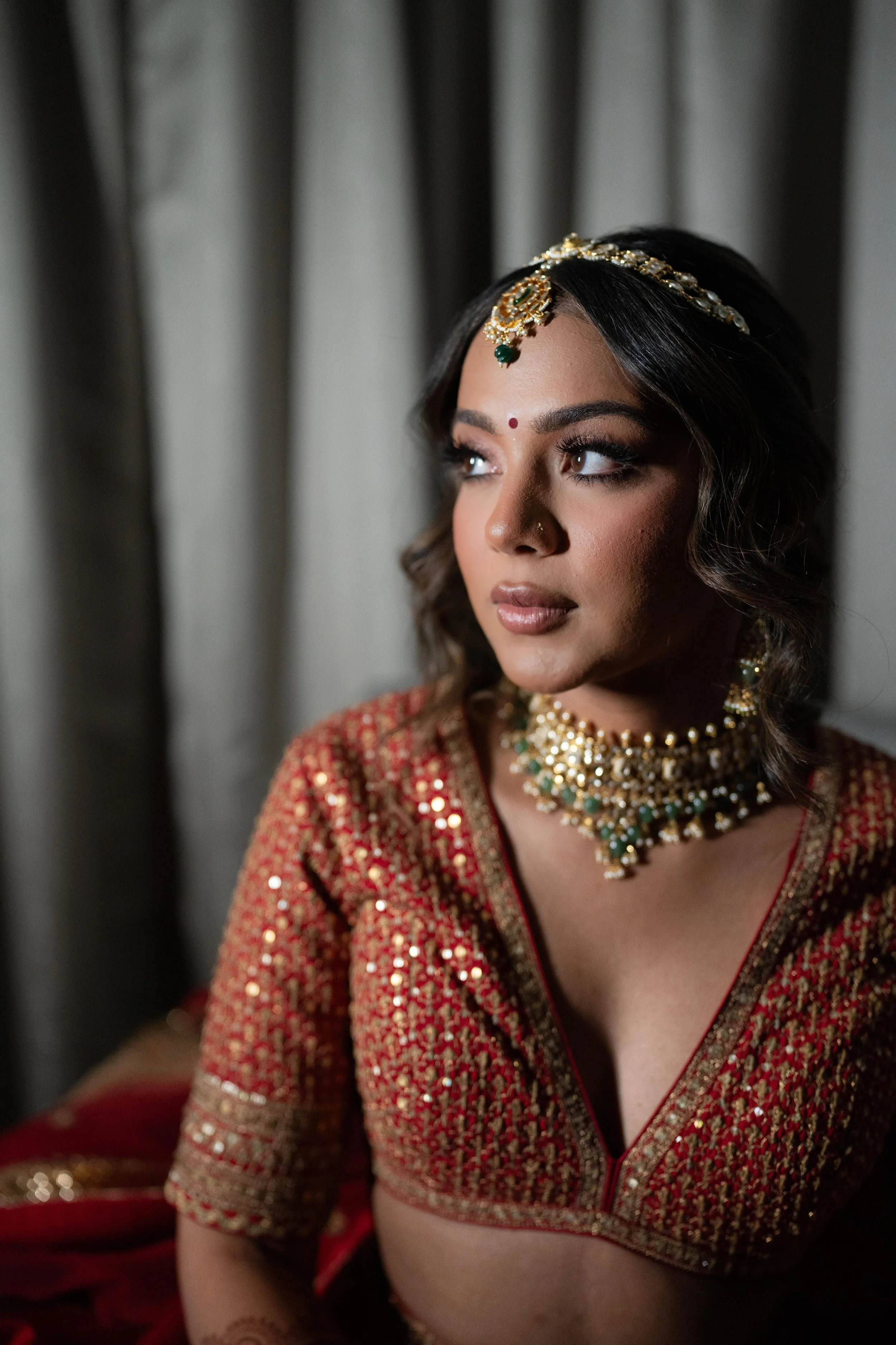 A bride dressed in traditional Indian attire with jewelry, looking slightly to the right, in front of curtains.